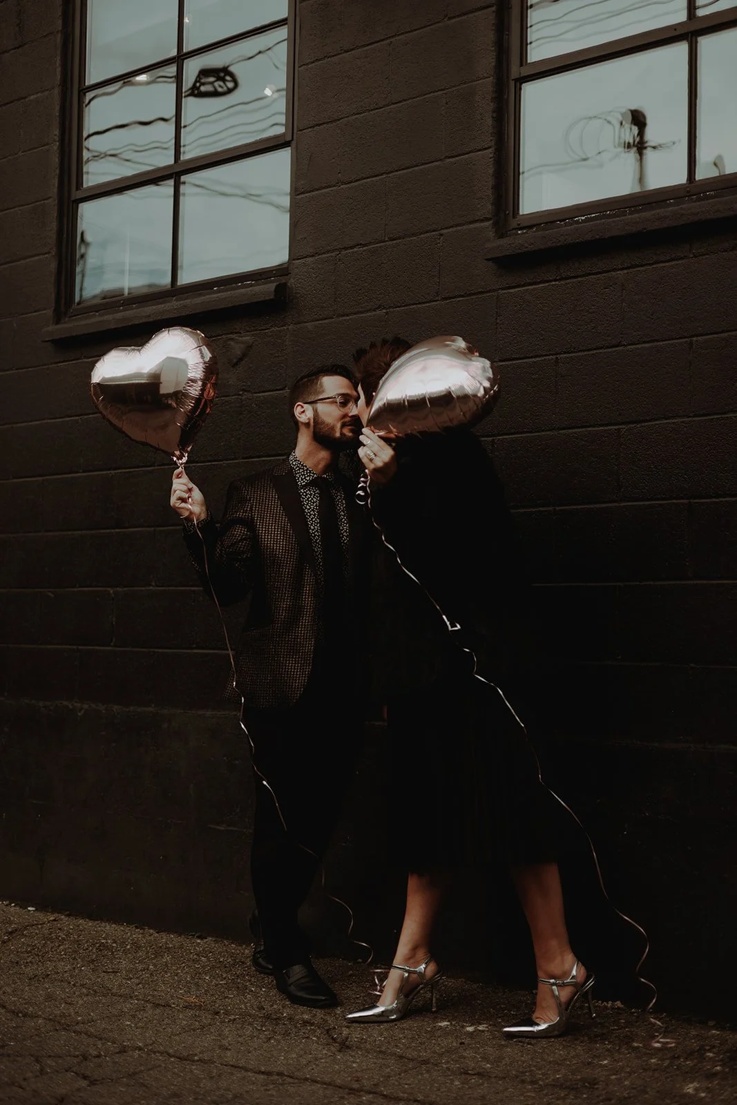 A man in a patterned jacket holding a heart-shaped balloon is leaning in to kiss a woman holding another heart-shaped balloon, standing against a dark brick wall with windows. They are dressed stylishly, and the scene has a romantic and intimate vibe