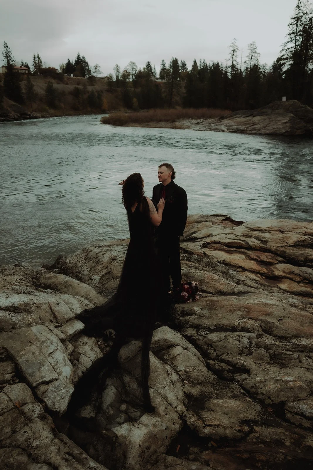 A couple stands on rocky riverbank, facing each other, with a river and forested landscape in the background. The woman wears a long black dress, and the man wears a dark suit.