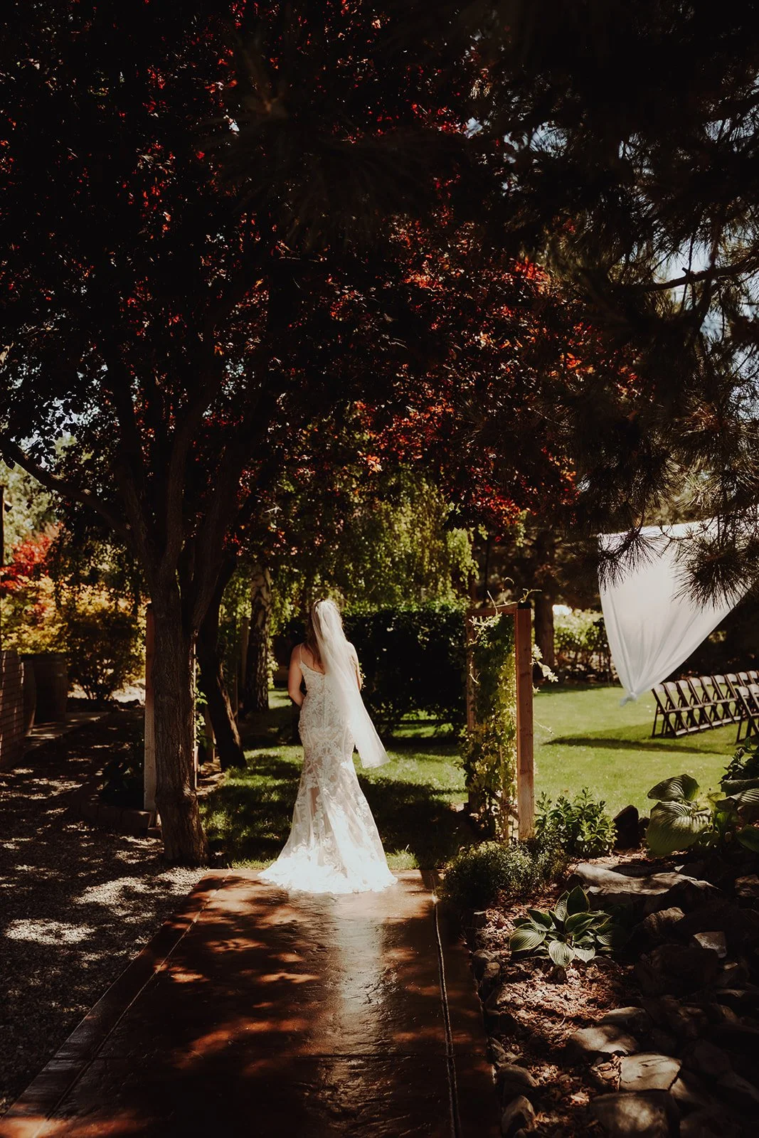 A bride in a lace wedding dress walking down a forested pathway, with trees and greenery on a sunny day.