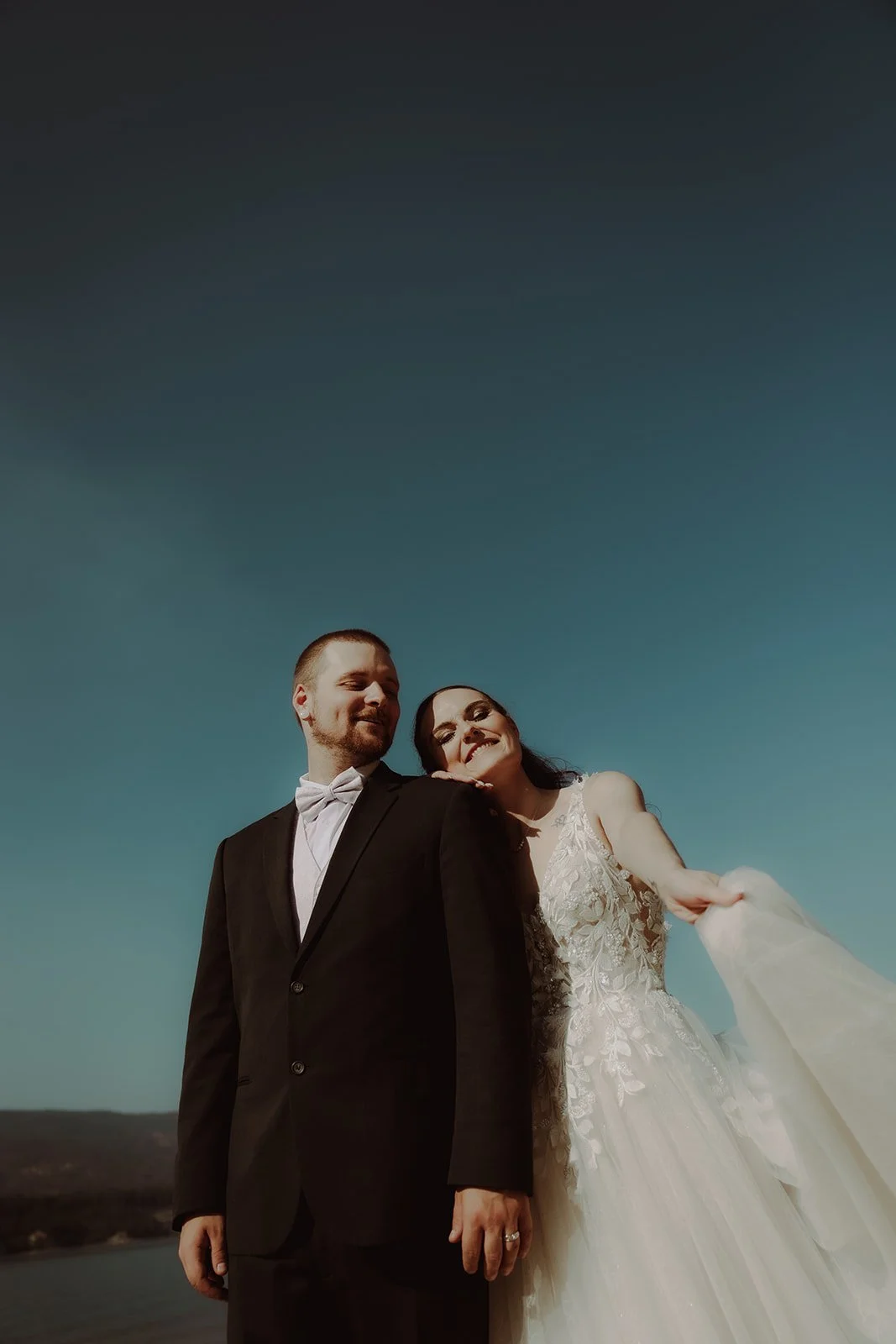 A bride and groom standing close outdoors under a blue sky, smiling, with the bride leaning her head on the groom's shoulder.