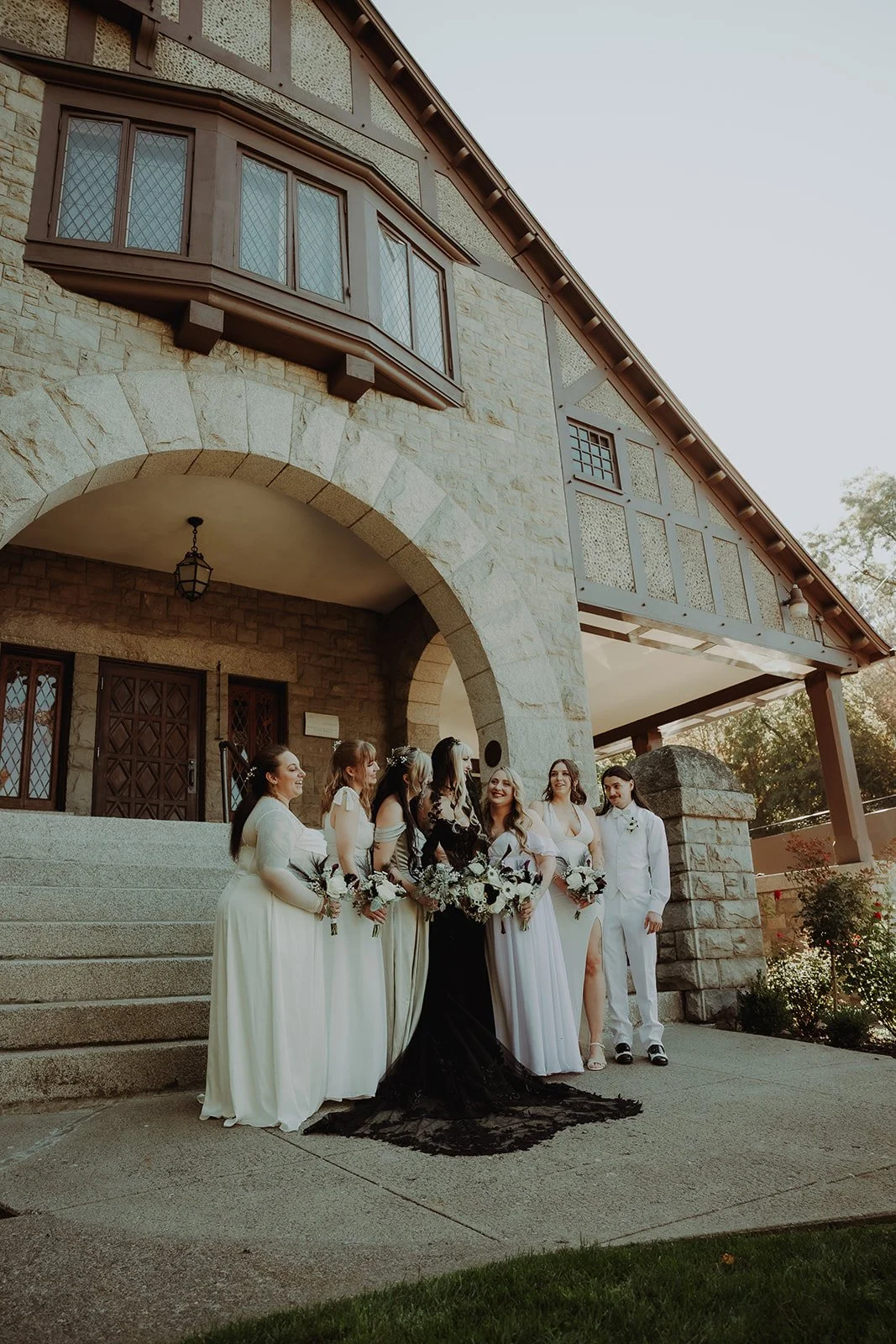 A group of women, including a bride in a black gown, and a groom in a white suit, stand outside a stone building with steps and an archway, smiling and holding bouquets, during a wedding celebration.