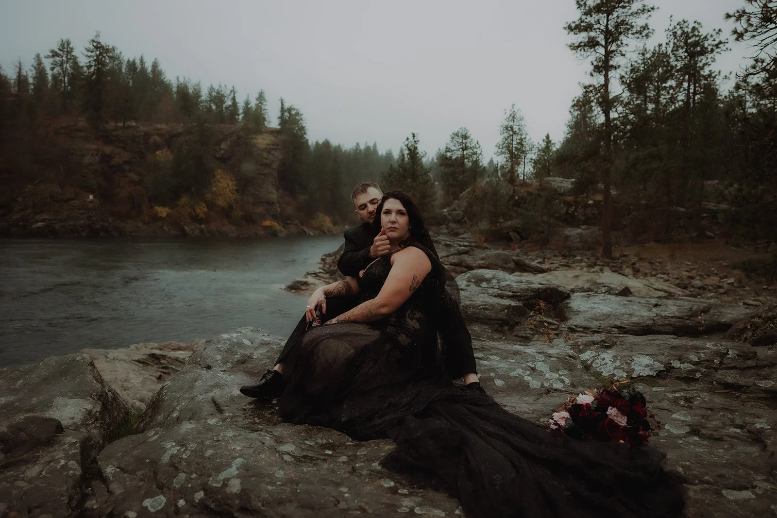 A couple dressed in black sitting on rocks by a river with trees in the background on a cloudy day.