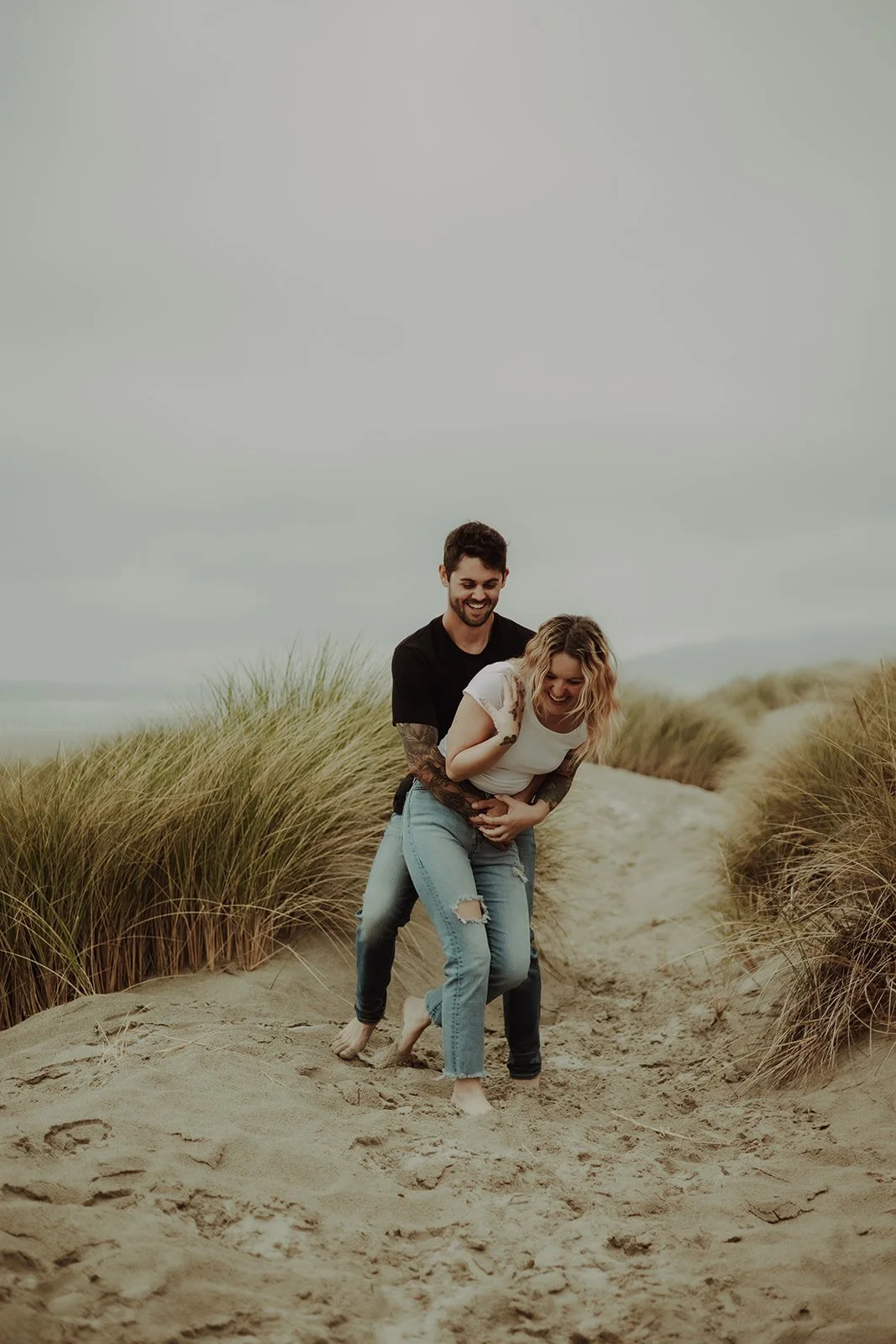A couple having fun on a sandy path with grassy dunes on either side, with an overcast sky above.