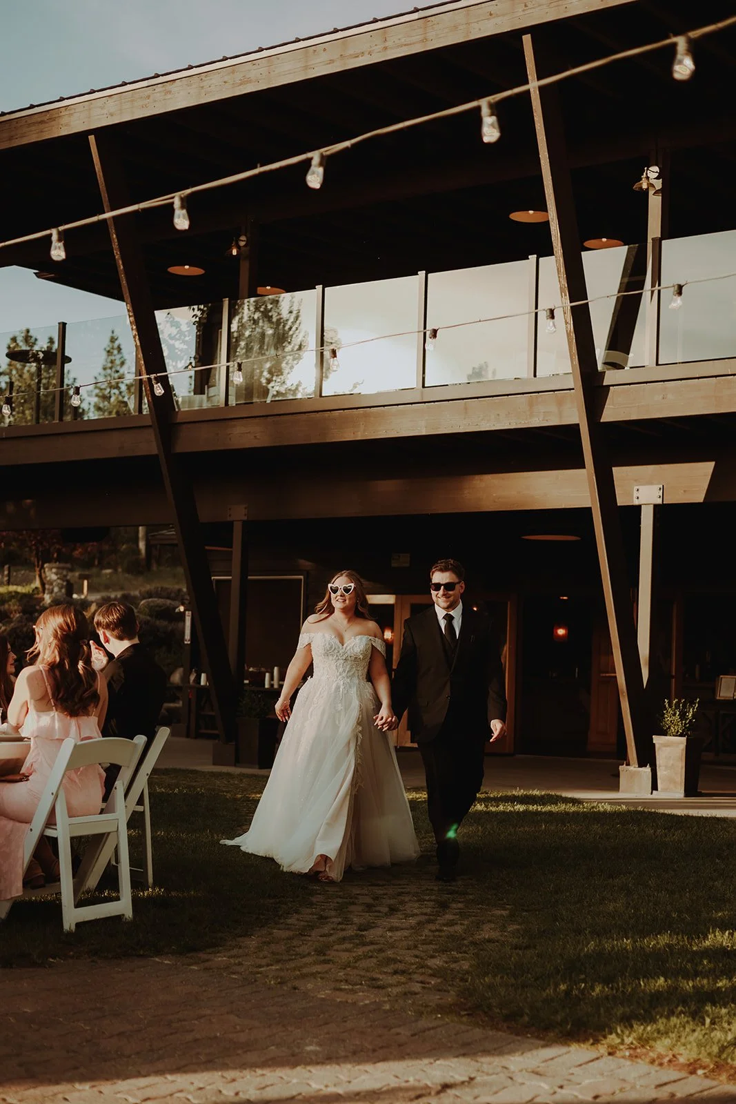 A newlywed couple walking hand in hand outside a modern building during sunset, with wedding guests sitting at tables nearby.