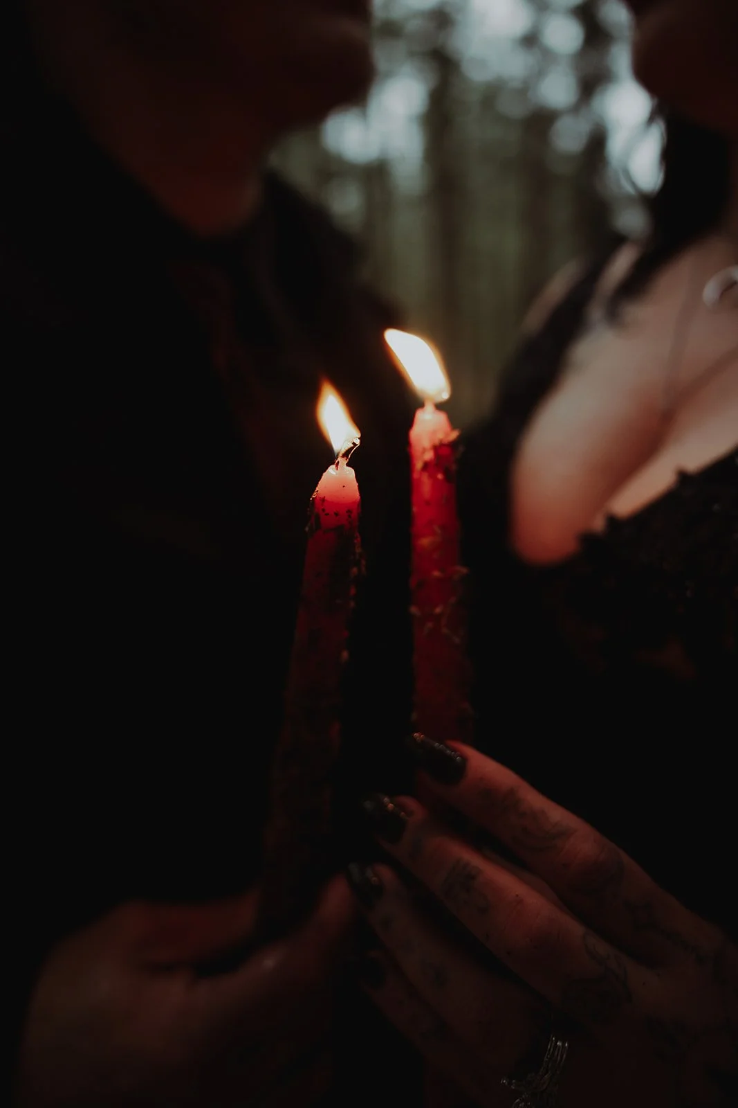Close-up of two pink birthday candles with lit flames being held by a person with tattooed hands. Dark background with blurred figures.