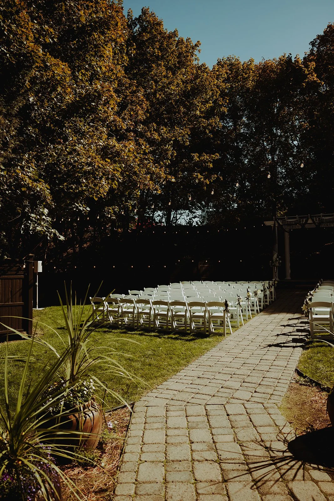 Outdoor wedding ceremony area with white chairs arranged along a brick aisle, surrounded by lush green grass and trees, under a clear blue sky.