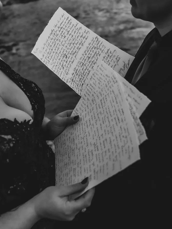 A person in a black dress holding handwritten letters, standing outdoors with another person visible in the background.