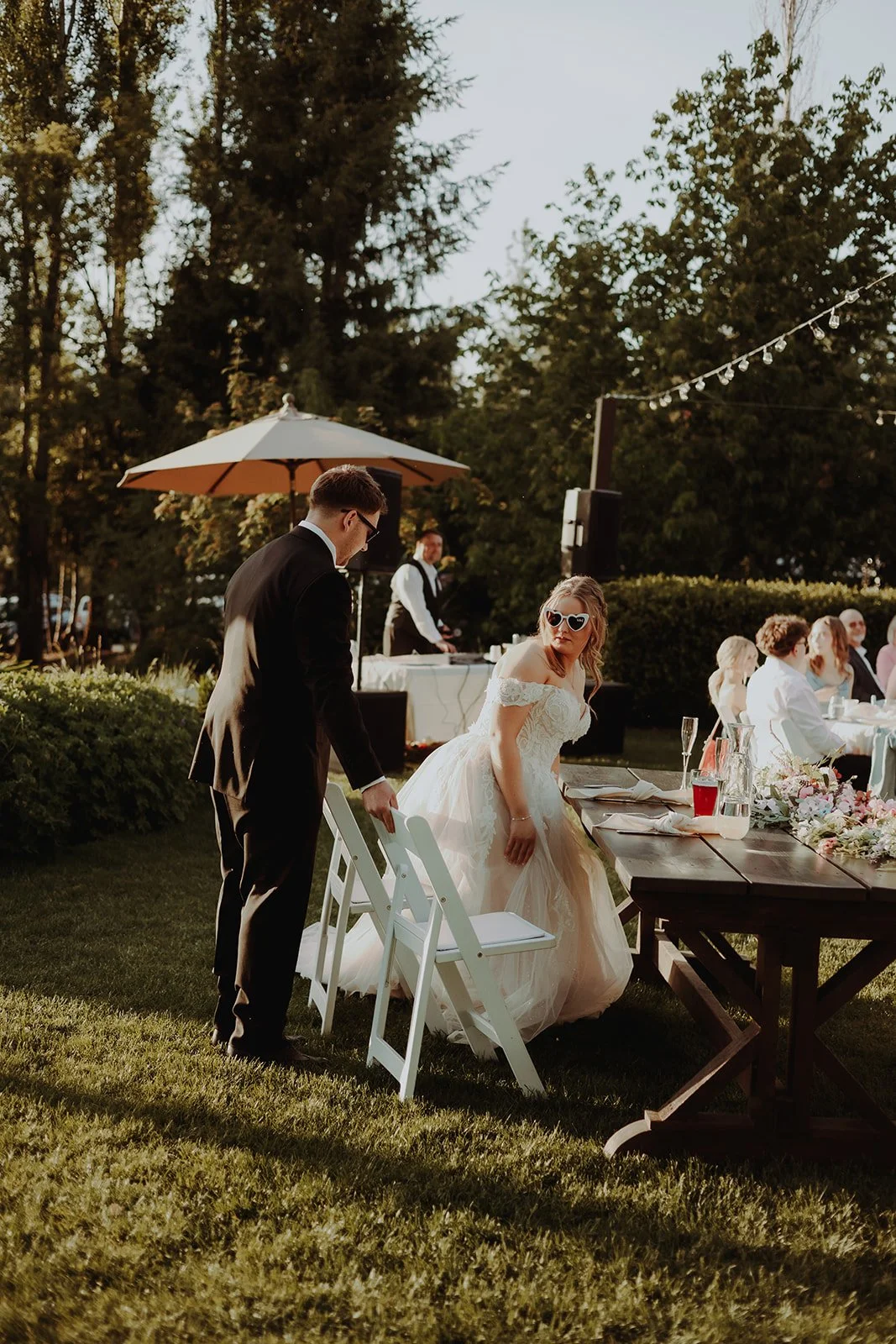 Bride in a wedding dress and sunglasses, and groom in a tuxedo, at an outdoor wedding reception during sunset, with guests seated at tables and someone DJing in the background.