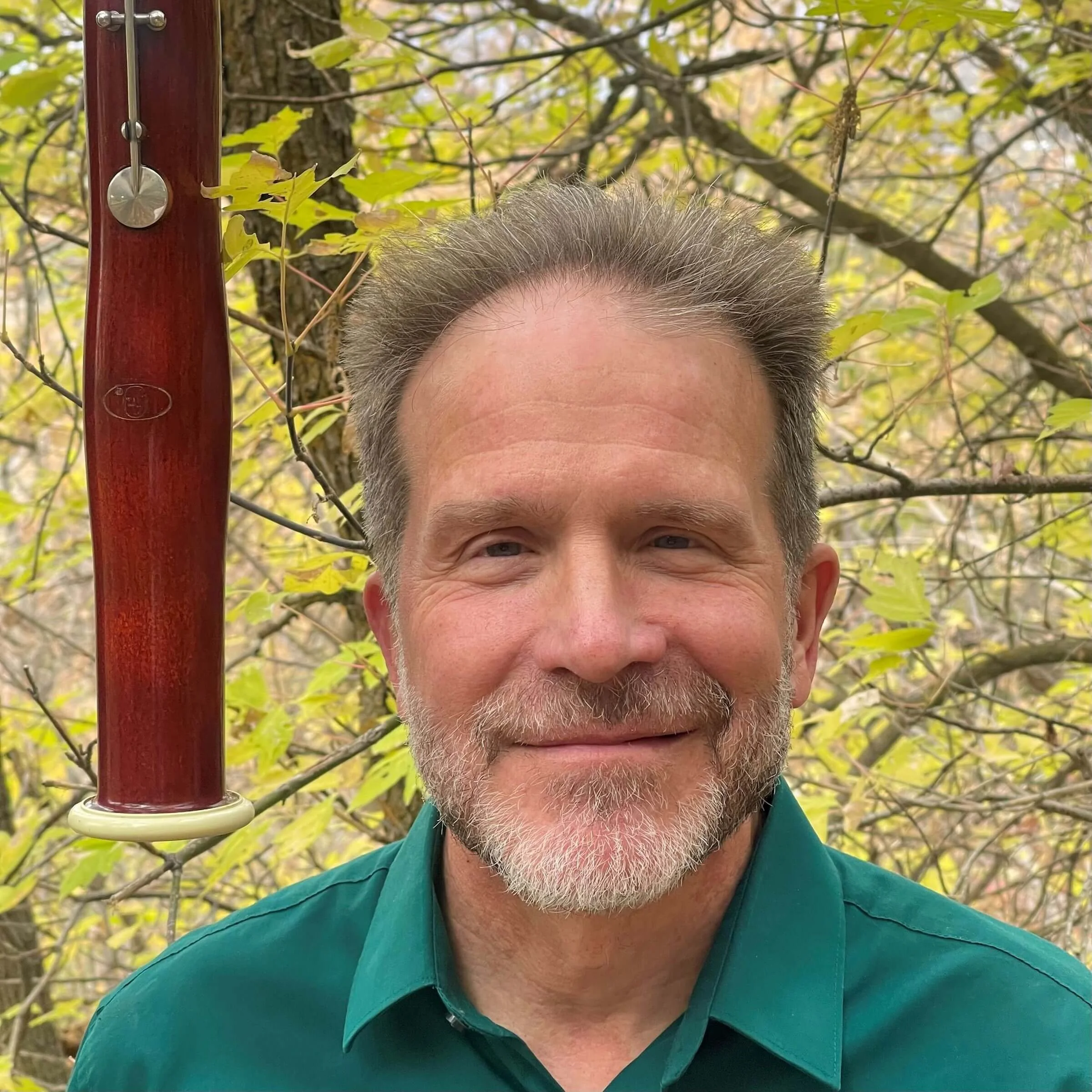 A gentleman in a green polo against a tree with light green leaves and the top of a bassoon in the corner of the image