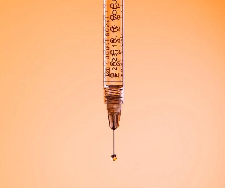 Close-up image of a medical syringe with a droplet at the needle's tip against a neutral peach background.