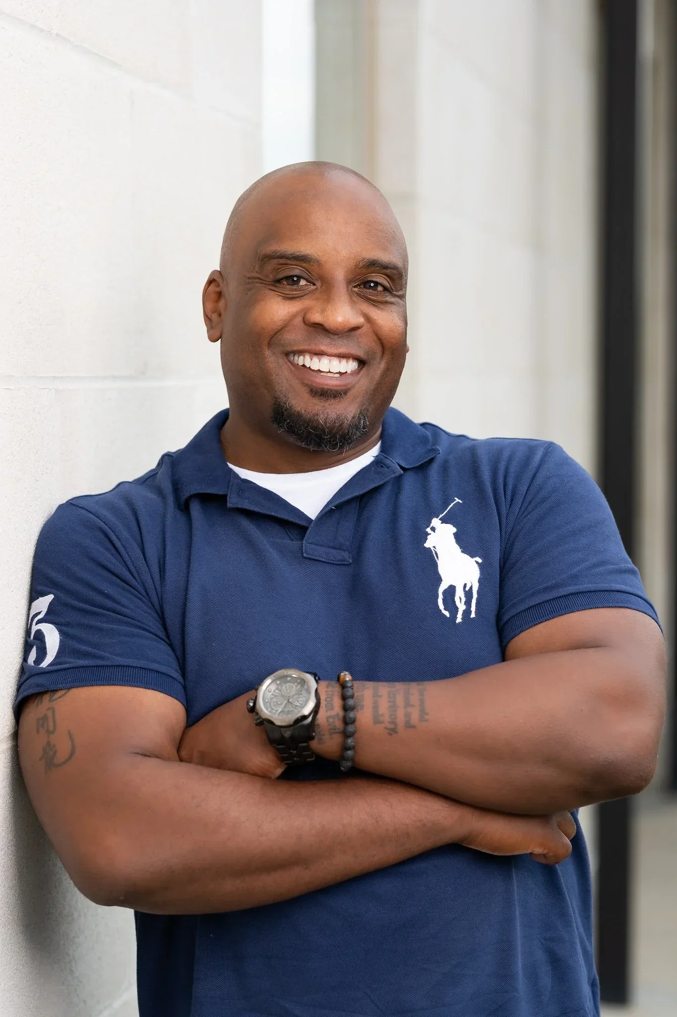 A smiling man with a beard and tattoos on his arms, wearing a blue polo shirt with a logo on it, standing against a light-colored wall with arms crossed.