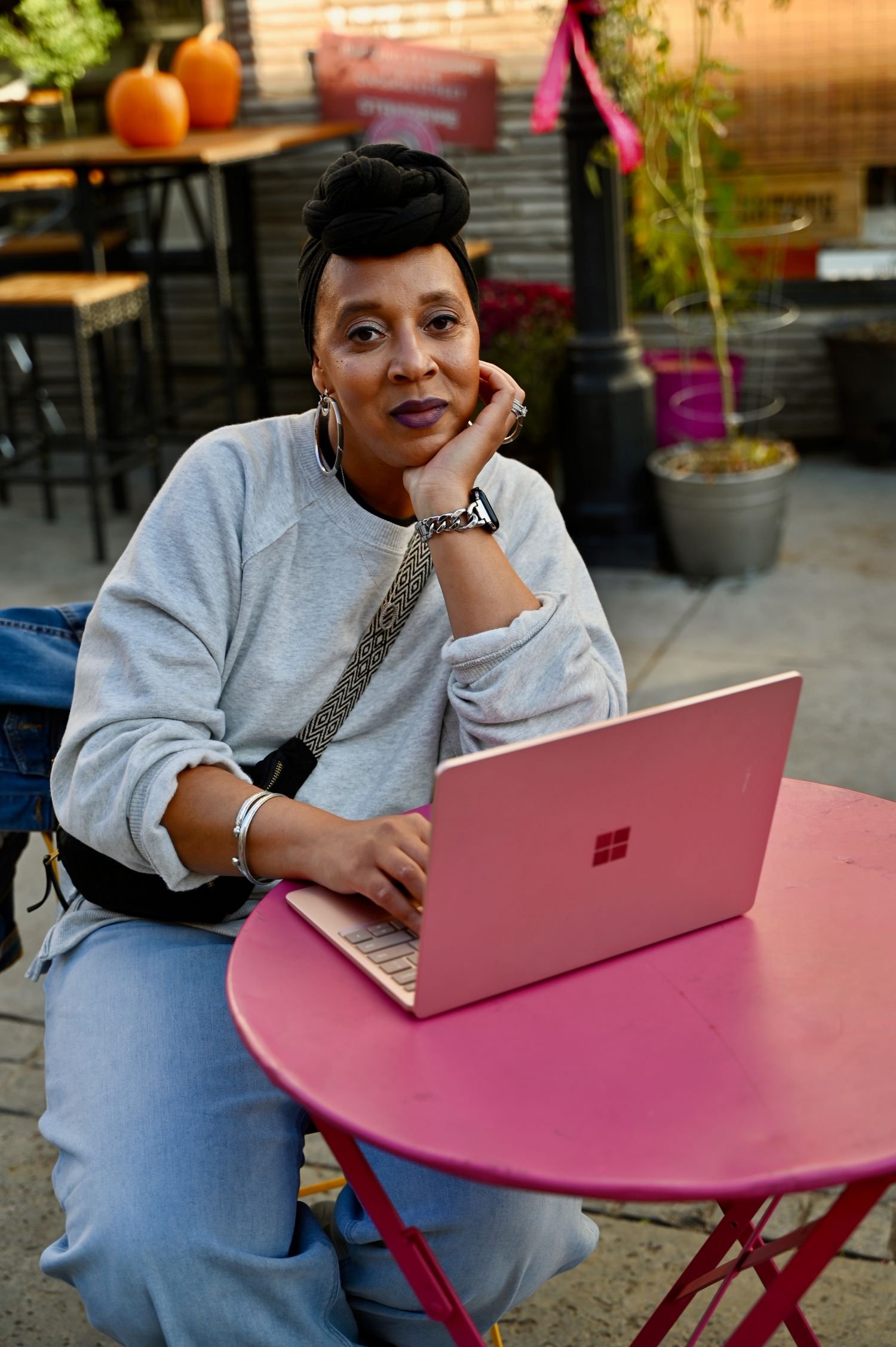A woman sitting at a pink outdoor table with a pink laptop, resting her chin on her hand, in an outdoor setting with autumn decor and plants in the background.