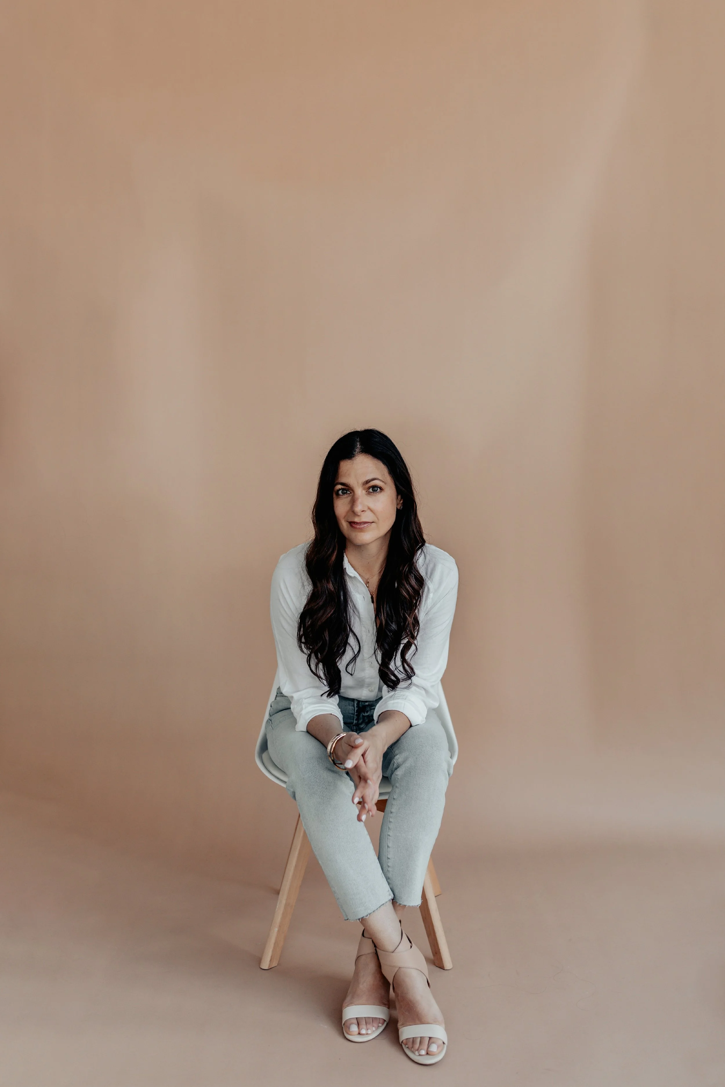 A woman with long dark hair, wearing a white shirt and light gray pants, sitting on a wooden stool against a plain beige background, with her legs crossed and her hands clasped.