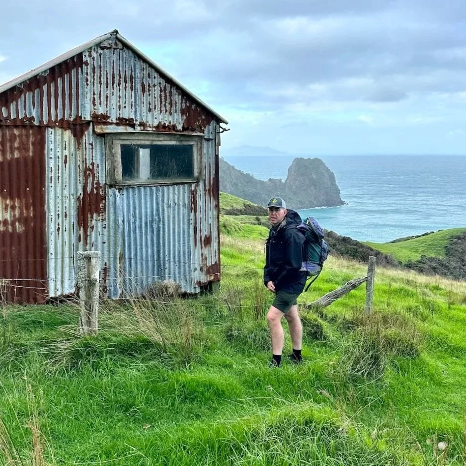 Andrew Gash standing on a grassy hillside beside a rusted corrugated iron shepherd's hut at Poley Bay on the Coromandel Walkway, with a steep rocky headland and open sea behind him, Aotea (Great Barrier Island) faintly visible on the horizon.