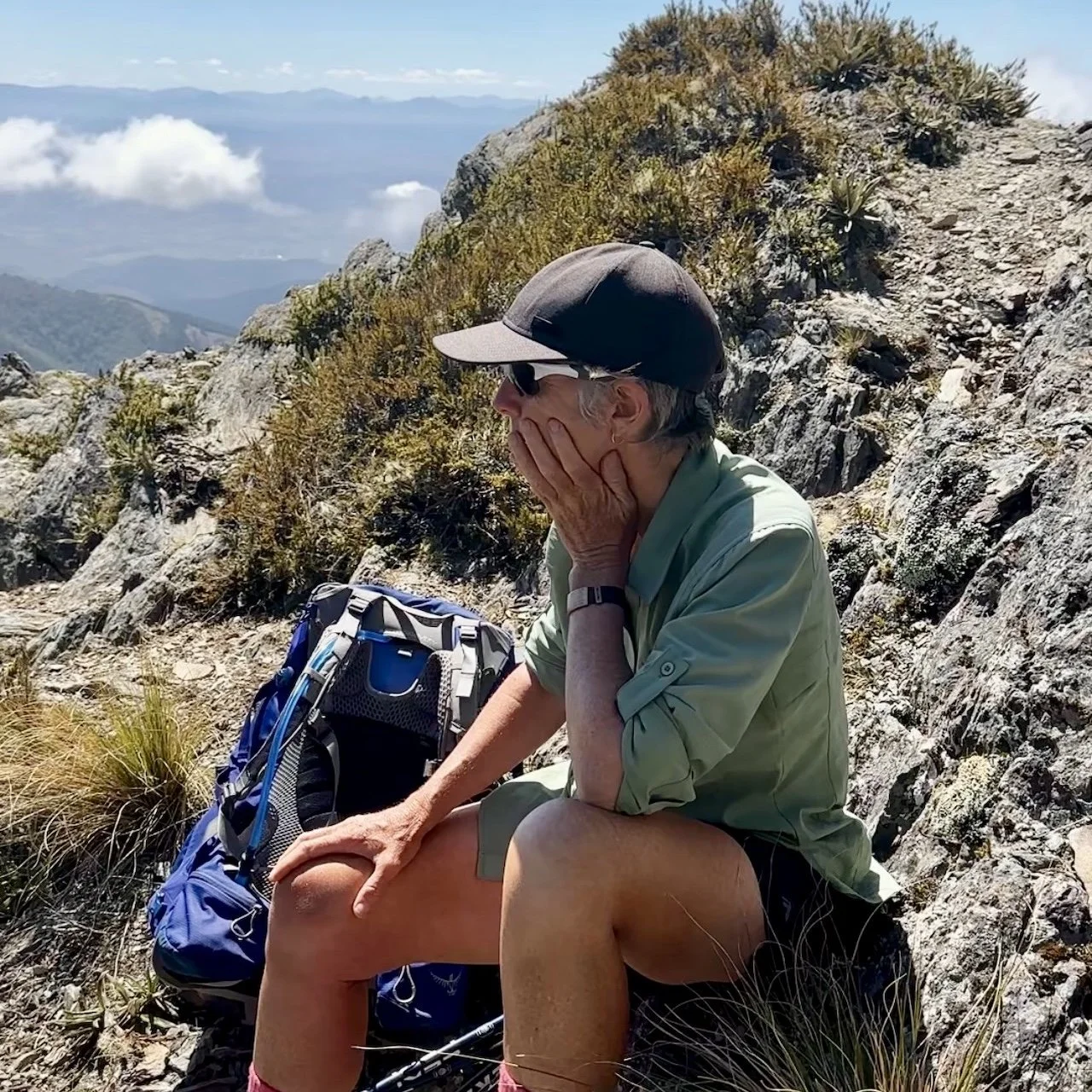 Sharon Evans sitting on rocky alpine terrain on the Richmond Range, Te Araroa, wearing a cap and daypack, hand to face in a moment of pause, with steep mountainside and distant valley visible behind her, New Zealand