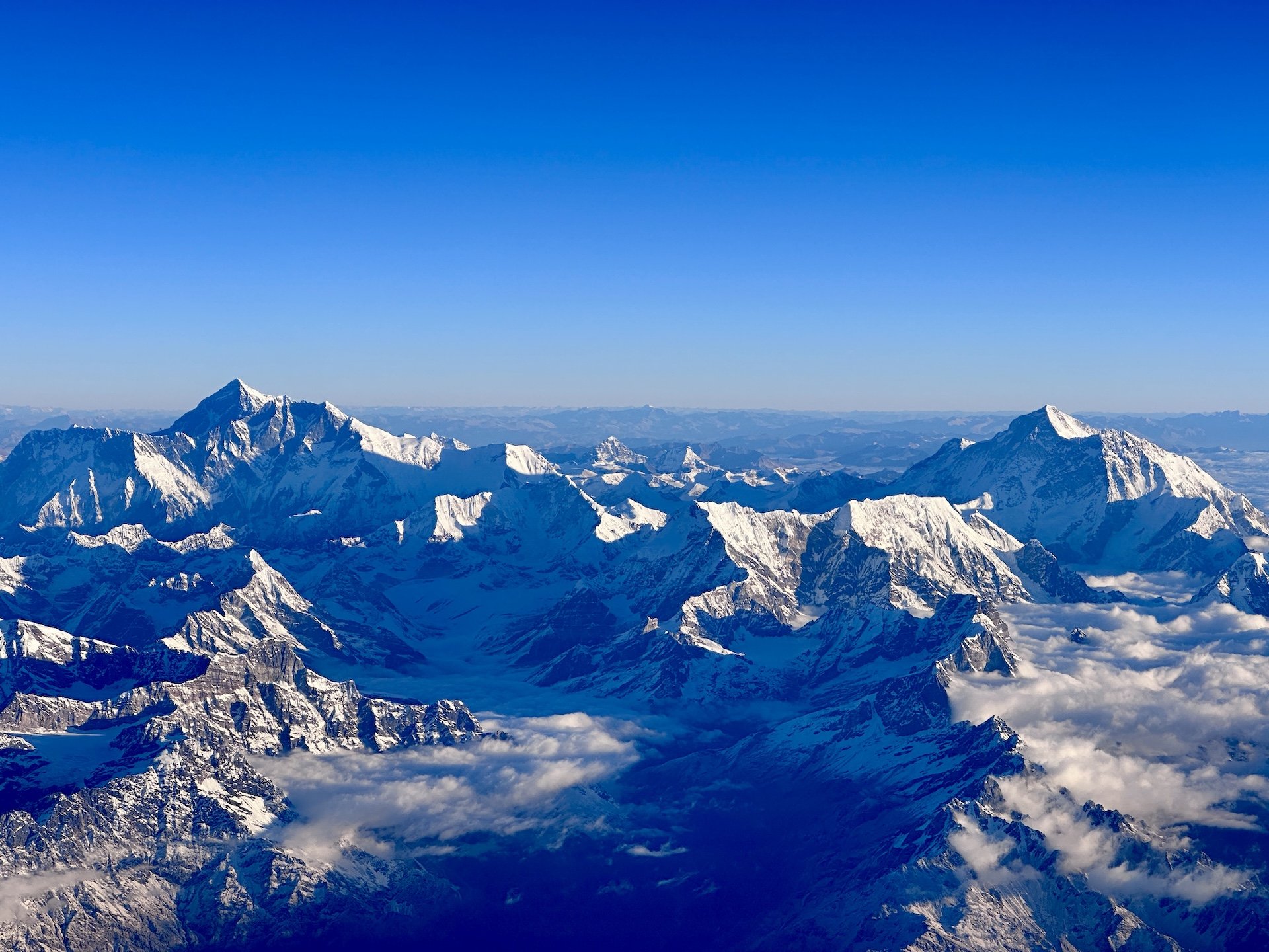 Aerial photograph of snow-covered Himalayan peaks with Mount Sagarmatha (Everest) at centre. Deep valleys, clouds in lower elevations, and surrounding snow-capped mountains visible under clear blue sky.
