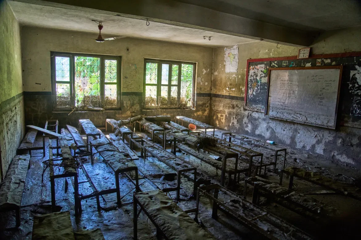 Flood-damaged classroom in Rasuwa district, Nepal, with overturned desks covered in mud, stained walls, and chalkboards visible. Windows show vegetation outside.
