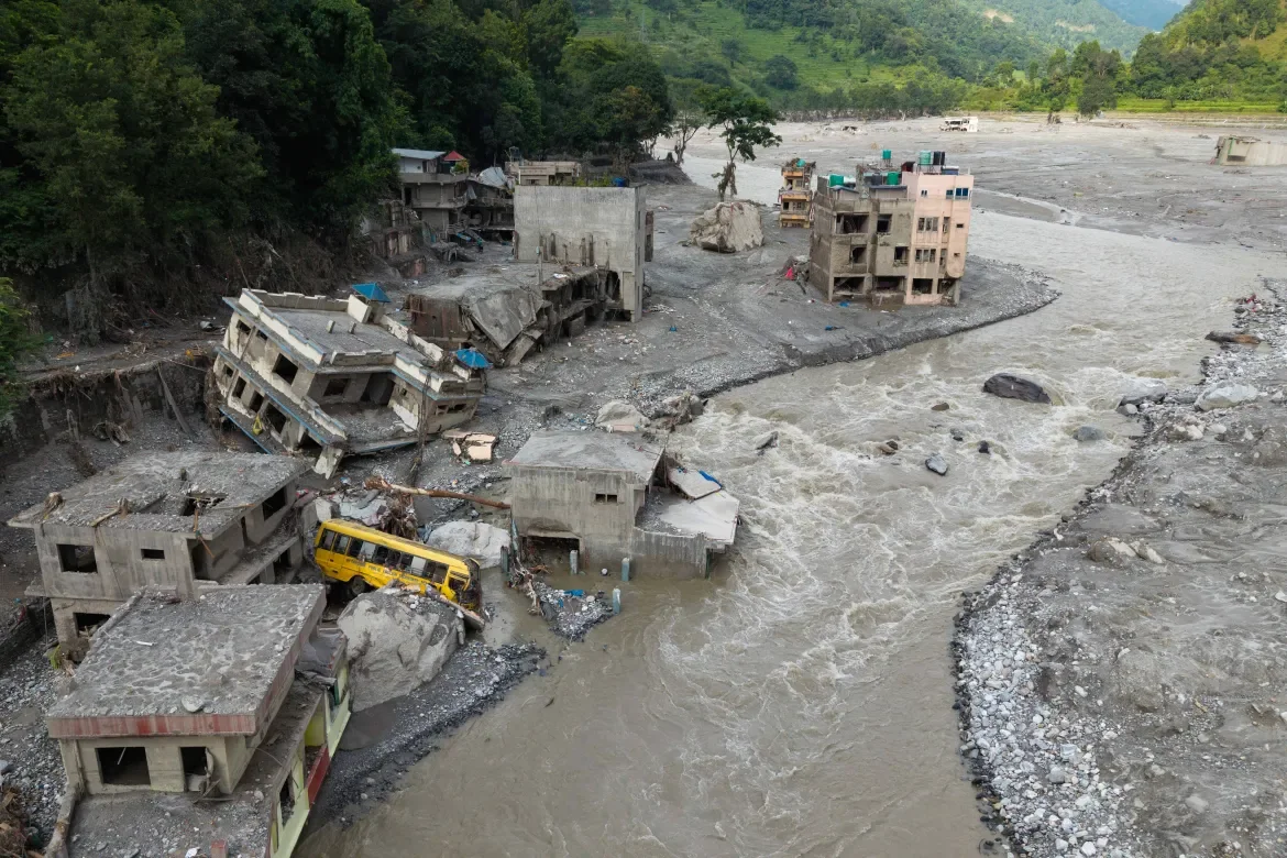 Aerial view of destruction along the Trishuli River in Betrawati, Nuwakot district. Concrete buildings damaged and partially collapsed, vehicles submerged in gray-brown muddy floodwaters. Mountains with green vegetation visible in background.