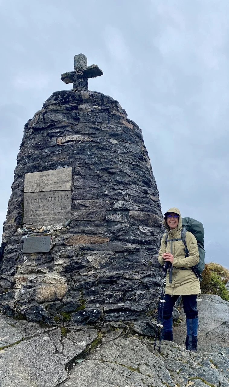 Hiker with backpack and trekking poles standing beside the stone cairn memorial to Quintin Mackinnon at the summit of Mackinnon Pass, the highest point on the Milford Track.