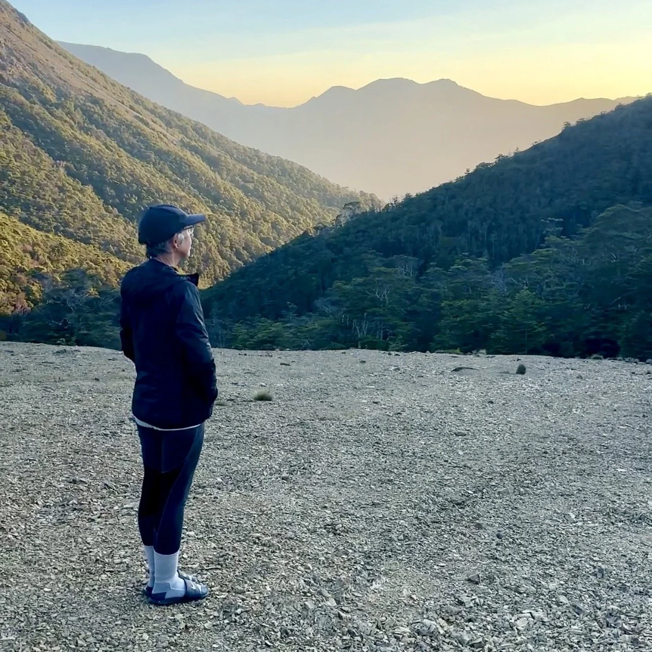 Sharon Evans standing on a scree plateau on the Richmond Range, Te Araroa, looking out toward Mount Richmond at sunset, with forested valleys and mountains in golden evening light, New Zealand.