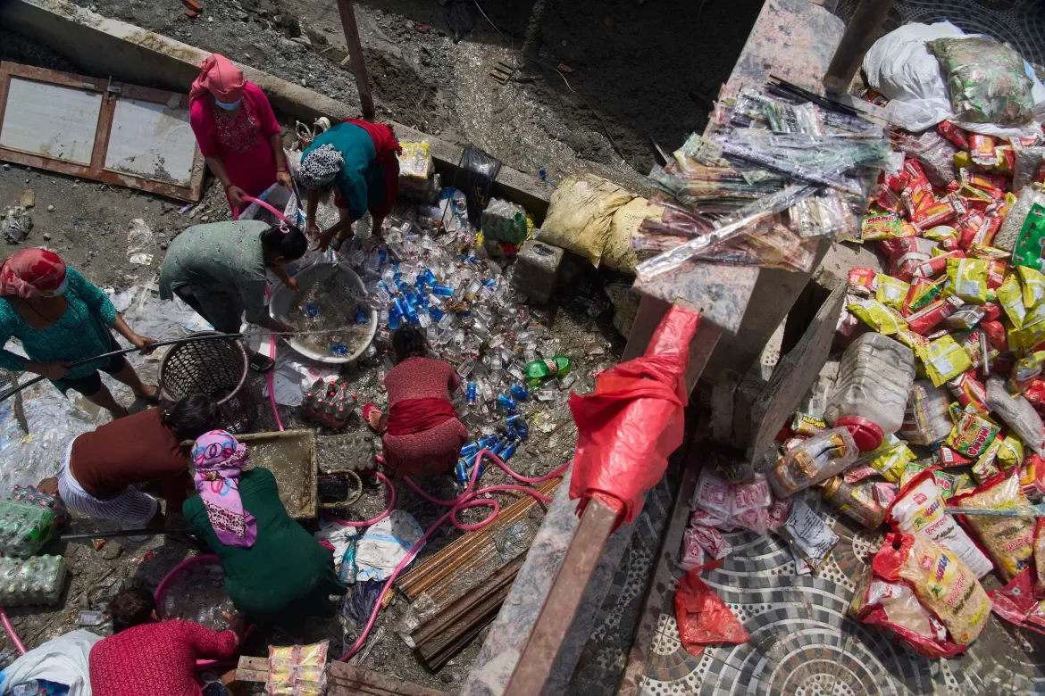 Overhead view of people salvaging goods from a flood-damaged grocery store in Dhading district. Scattered packaged foods, household items, and debris cover the ground as people work to recover what can be saved.