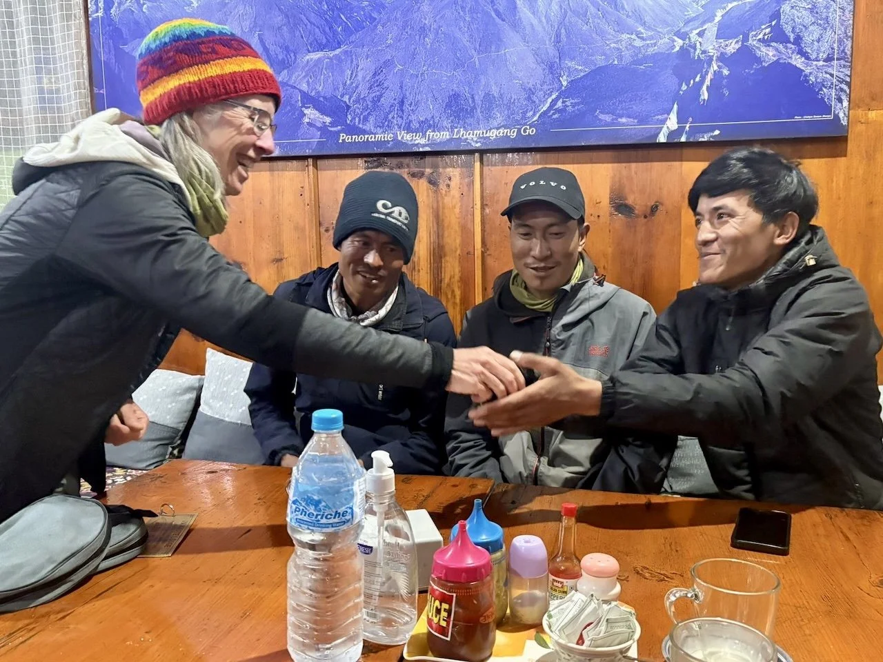 Sharon Evans laughing with three Nepali porters at a wooden table in Lukla. They're doing a group hand stack gesture. Everest Base Camp panoramic map visible on wall behind them. Water bottles and condiments on table.