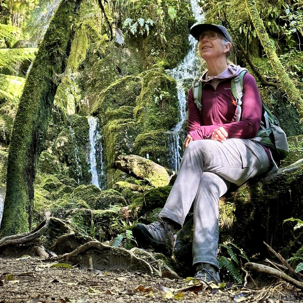 Sharon Evans seated on a mossy tree root at the base of Cascade Falls, Te Auheke Track, Ōkataina Trails, Bay of Plenty, surrounded by native ferns and moss-covered rocks with waterfalls cascading behind her.
