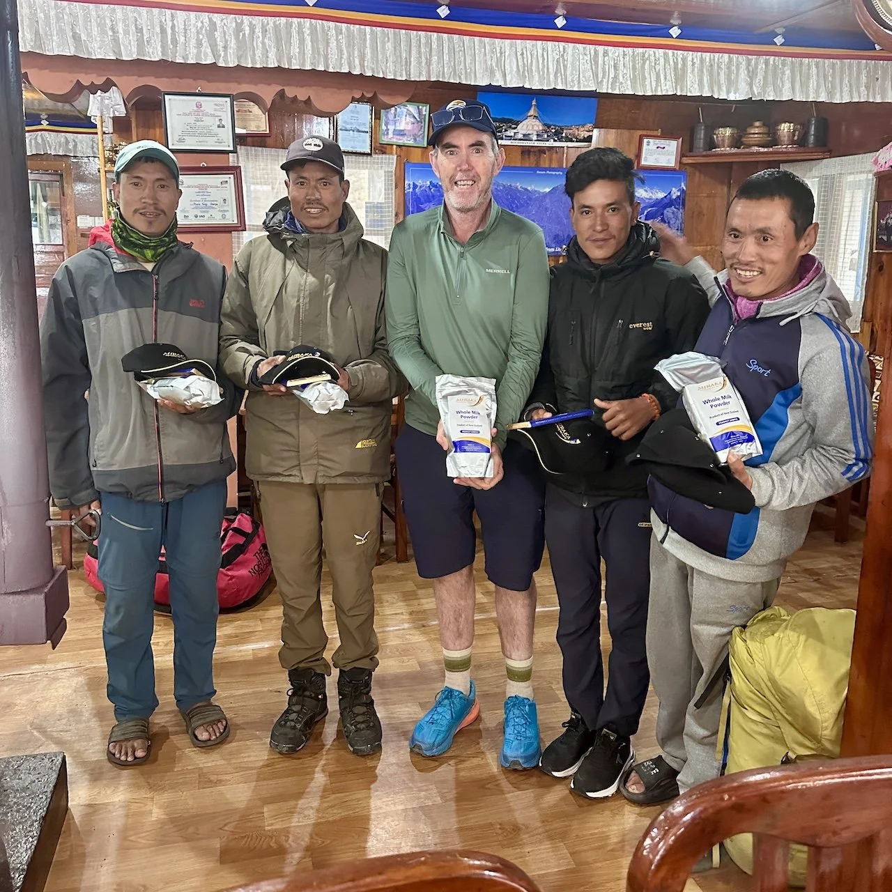 Andrew Gash standing with four Nepali porters in Lukla lodge, all holding supplies. Mountain photographs on walls behind them. Indoor trekking lodge setting.