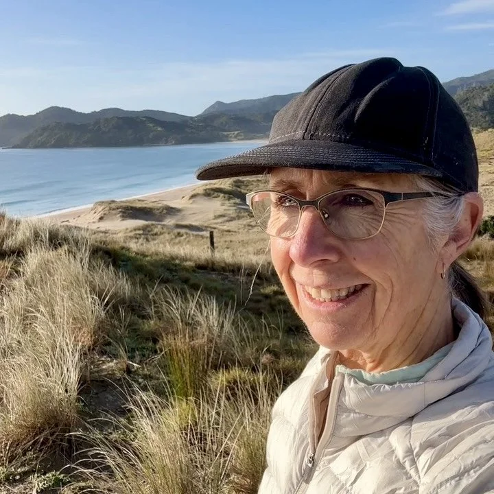 Sharon Evans smiling at the camera wearing a black cap and glasses, with Waikawau Beach and native bush-covered hills visible behind her, Coromandel Peninsula, New Zealand.