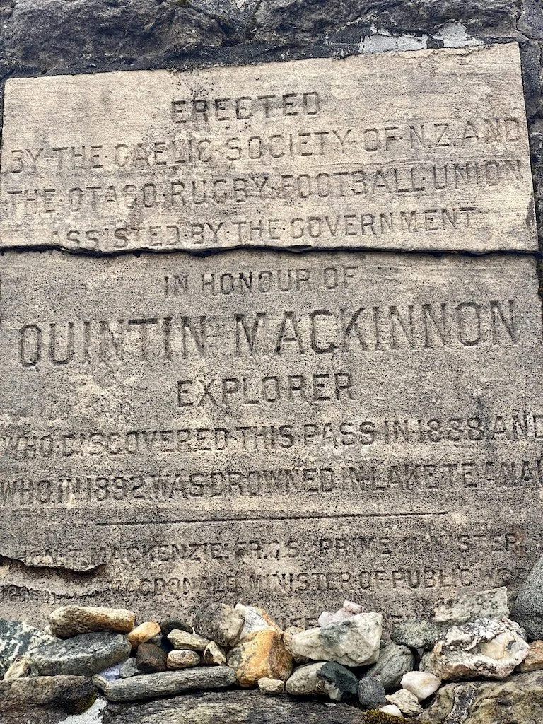 Weathered stone plaque on the Milford Track commemorating explorer Quintin Mackinnon, who discovered Mackinnon Pass in 1888, surrounded by river stones.