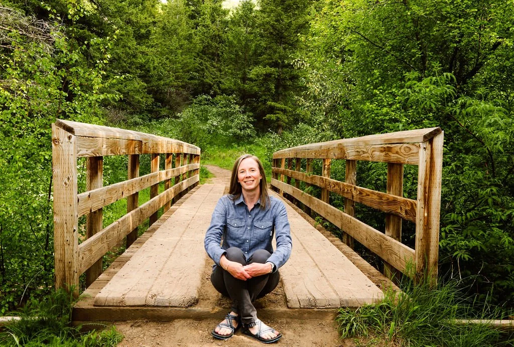 A woman sitting cross-legged on a small wooden bridge in a lush green forest, smiling at the camera.