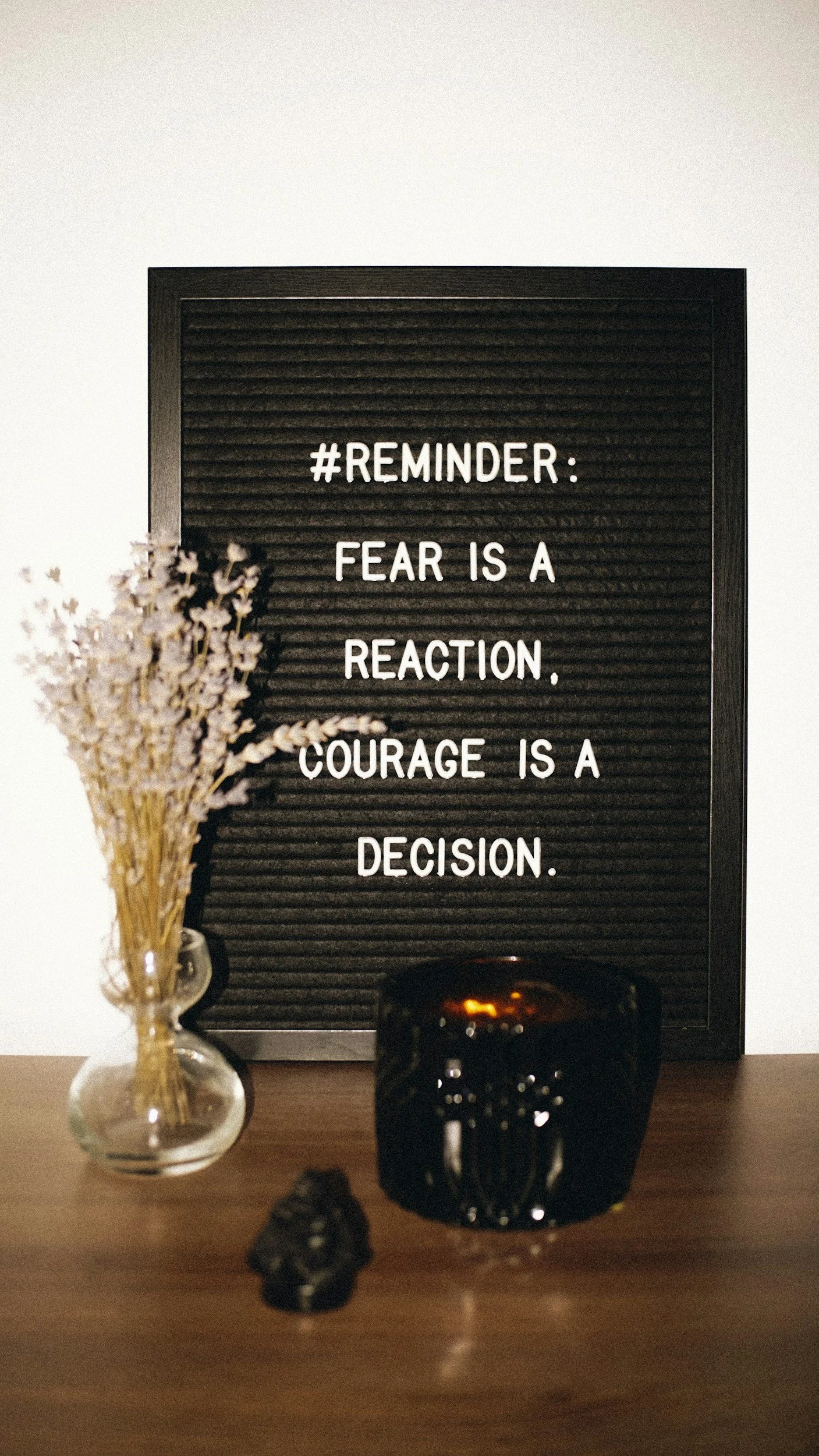 A black letter board with motivational quote, a glass vase with dried flowers, a dark candle holder, and a small black decorative object on a wooden surface.