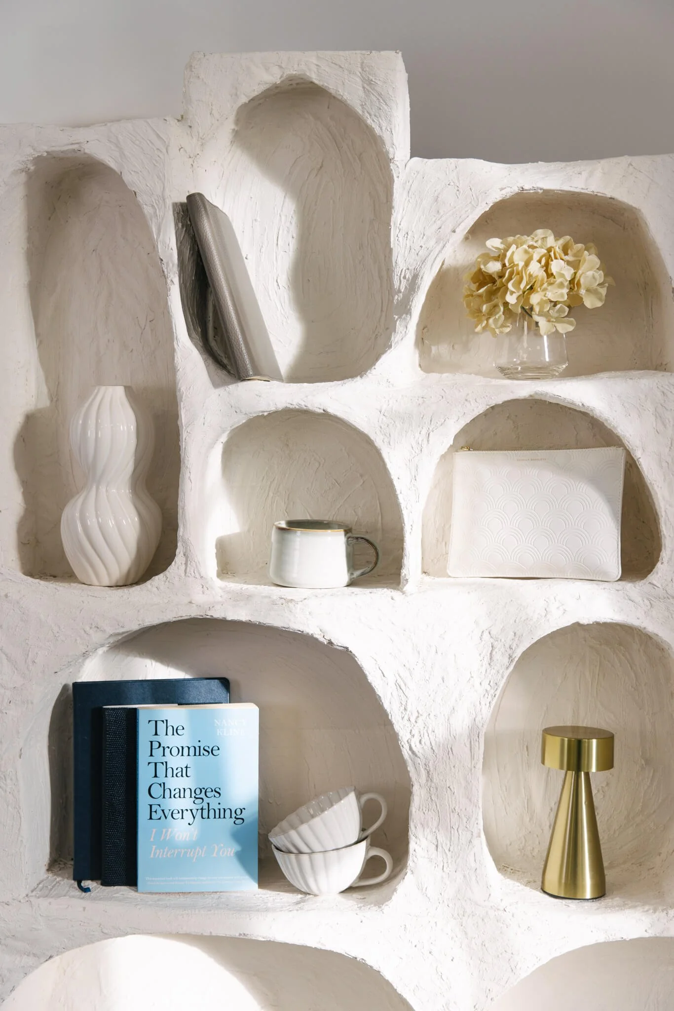 Decorative white wall shelf with various objects: a long gray item, a white ceramic vase, a bouquet of light yellow flowers in a glass bowl, a white textured bag, a ceramic mug, a hardcover book titled 'The Promise That Changes Everything' in blue and black, two white cups stacked, and a gold metallic vase.