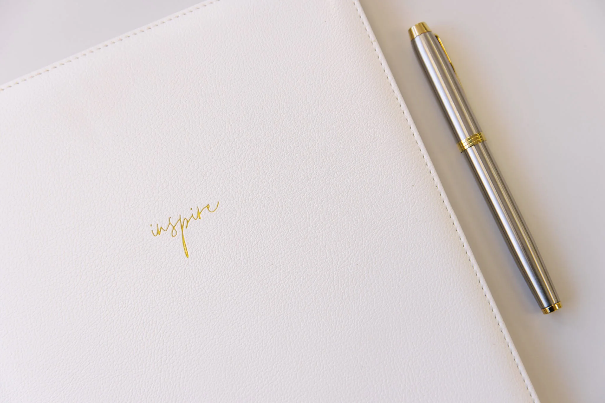 A white notebook with the word 'inspire' written in gold on the cover, placed next to a silver and gold pen on a white surface.