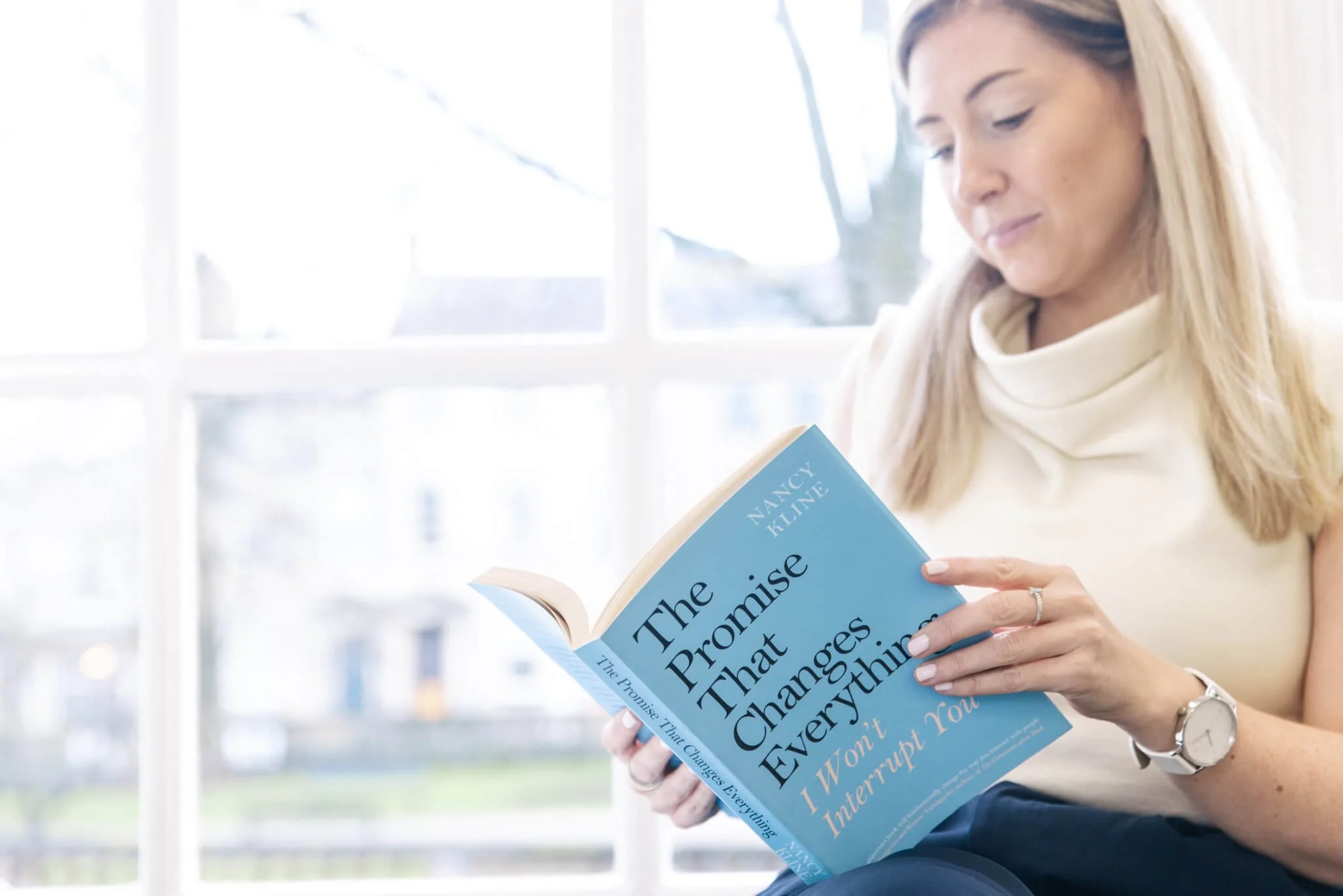 A woman sitting indoors by a large window, reading a book titled 'The Promise That Changes Everything' by Nancy Kline.