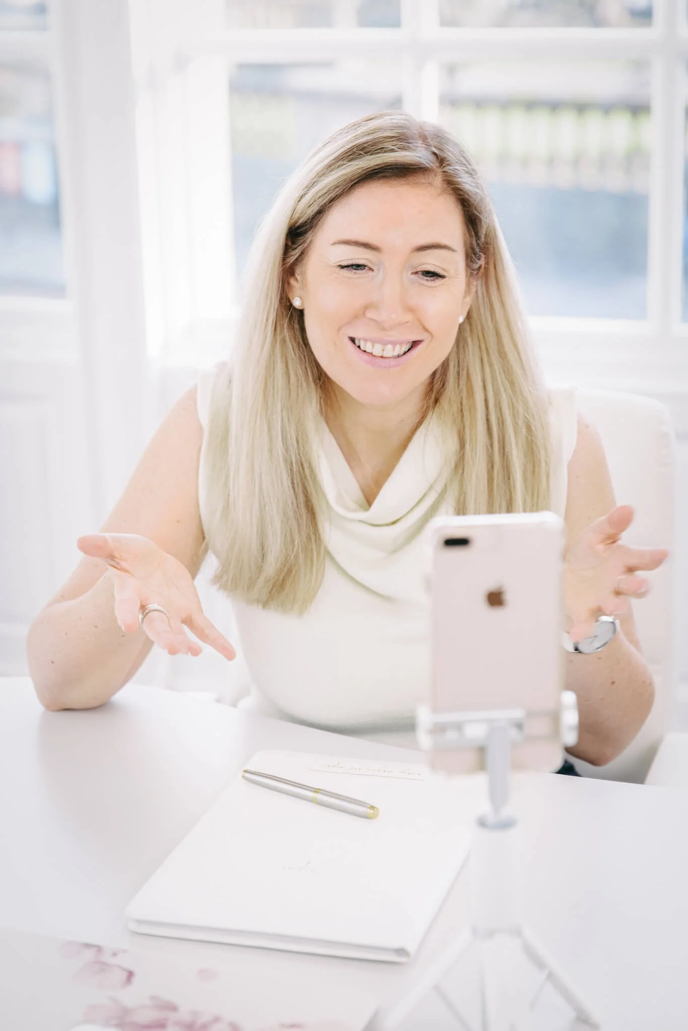 A woman with blonde hair and pearl earrings smiling and gesturing with her hands while looking at her phone, which is mounted on a stand on a white table with a notebook and pen, in a bright room with large windows.