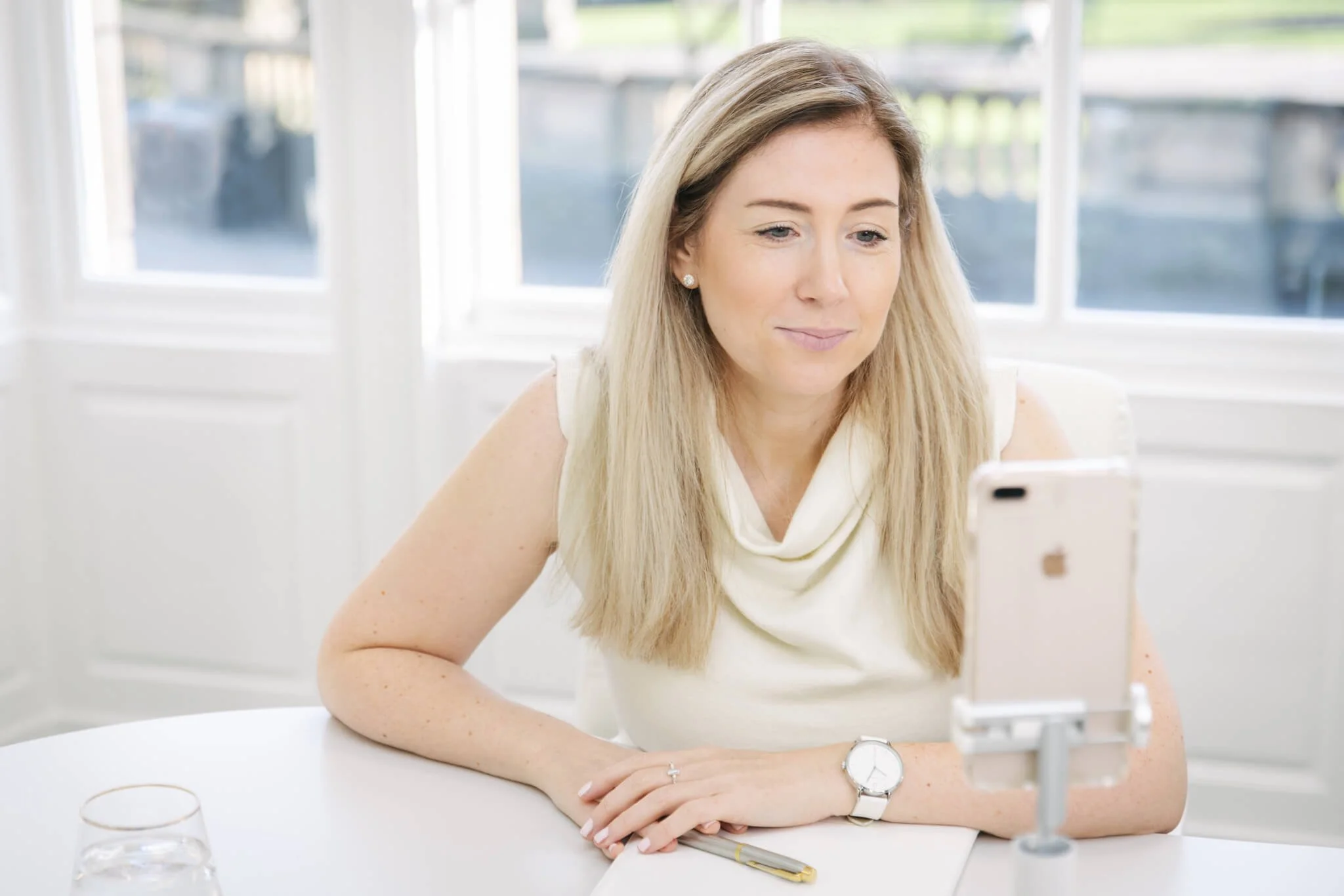 A woman with long blonde hair is sitting at a white table, looking at a smartphone on a stand. She is wearing a white sleeveless top, a watch, and earrings, with a notebook and a pen in front of her, and a glass of water nearby.