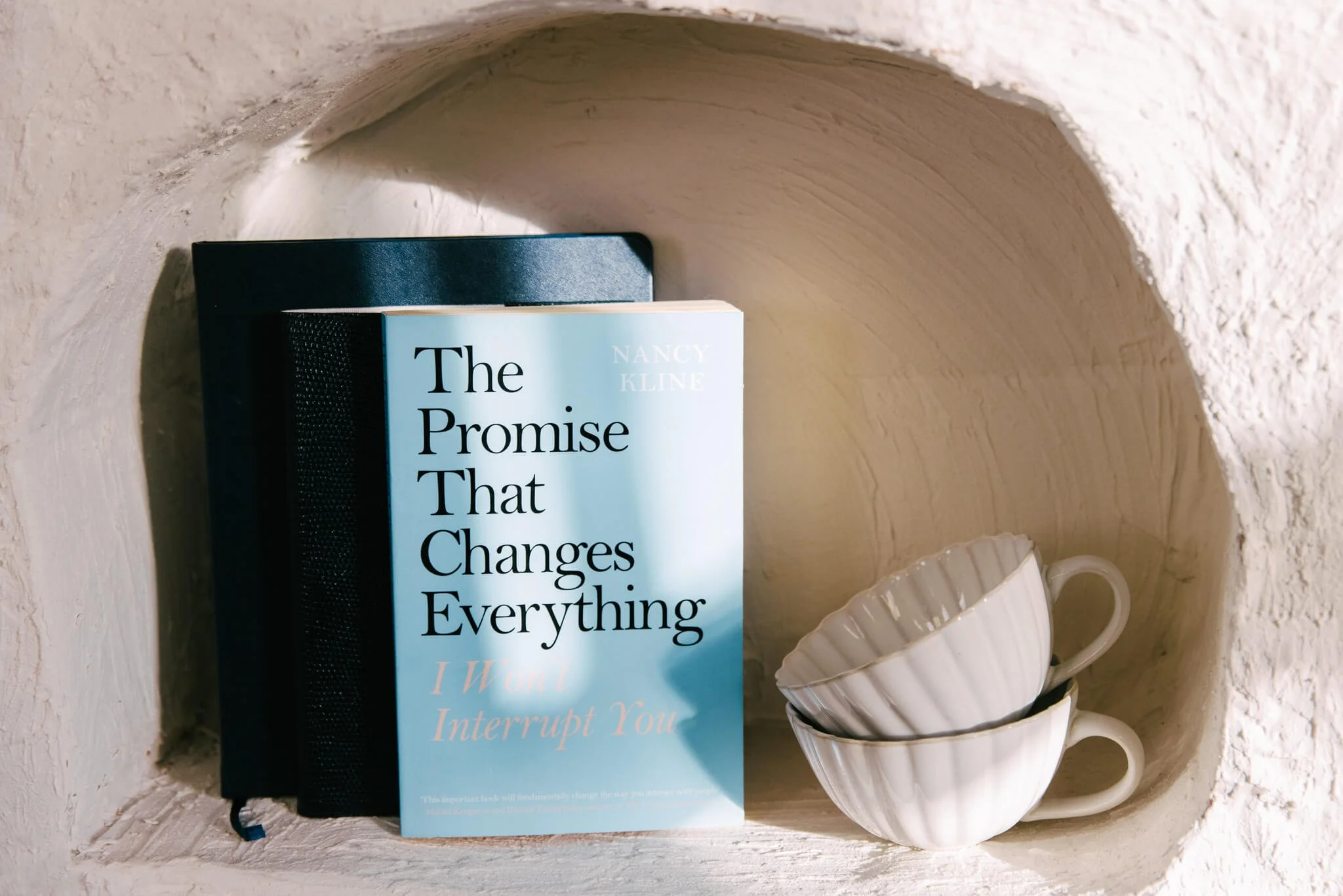 A bookshelf niche contains two stacked white teacups with a book titled 'The Promise That Changes Everything' by Nancy Kline and a closed black notebook or journal.