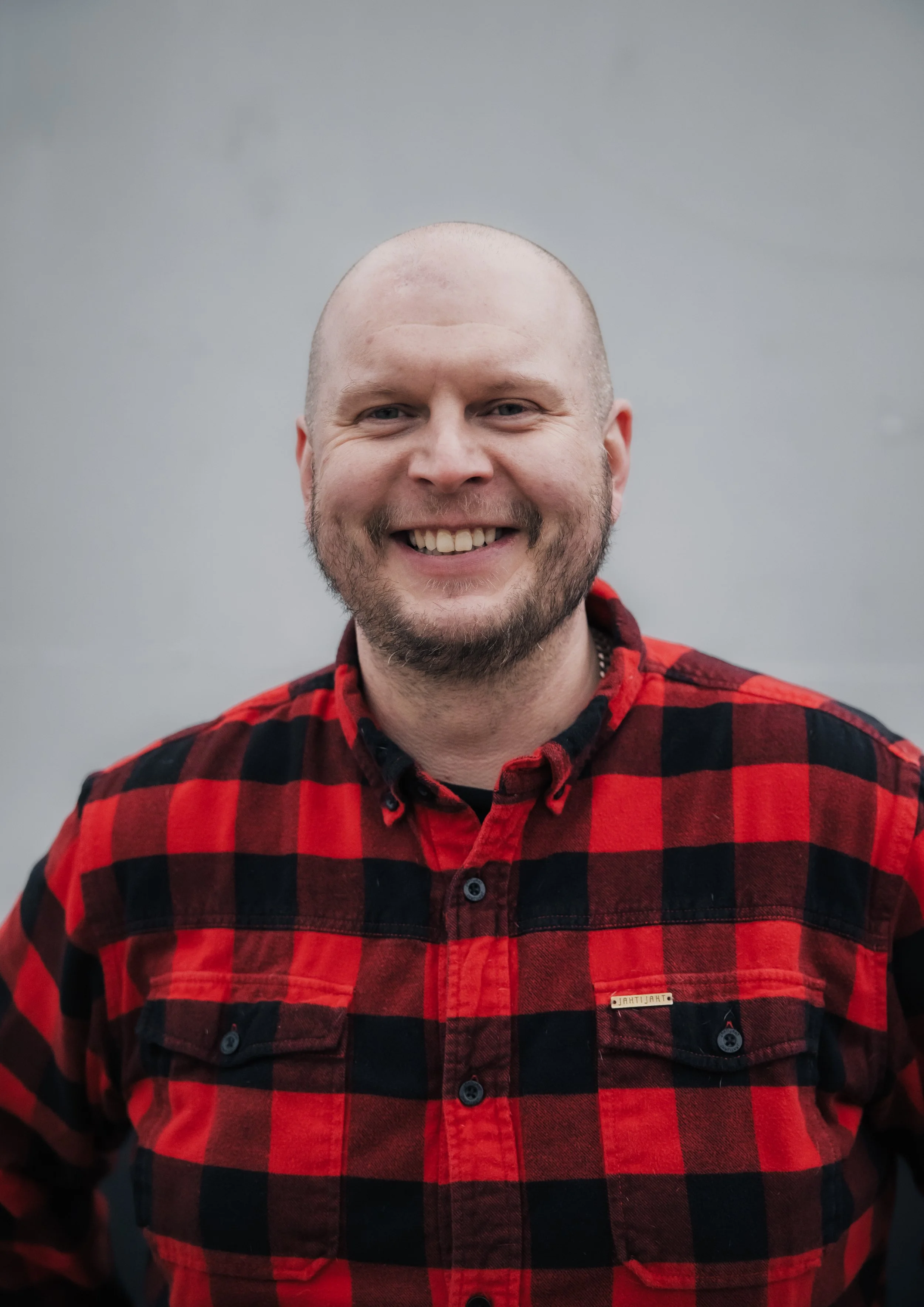 Hymyilevä henkilö, kokki ja yrittäjä Veli-Matti Nousiainen pukeutuneena punaiseen ruutupaitaan harmaata taustaa vasten.