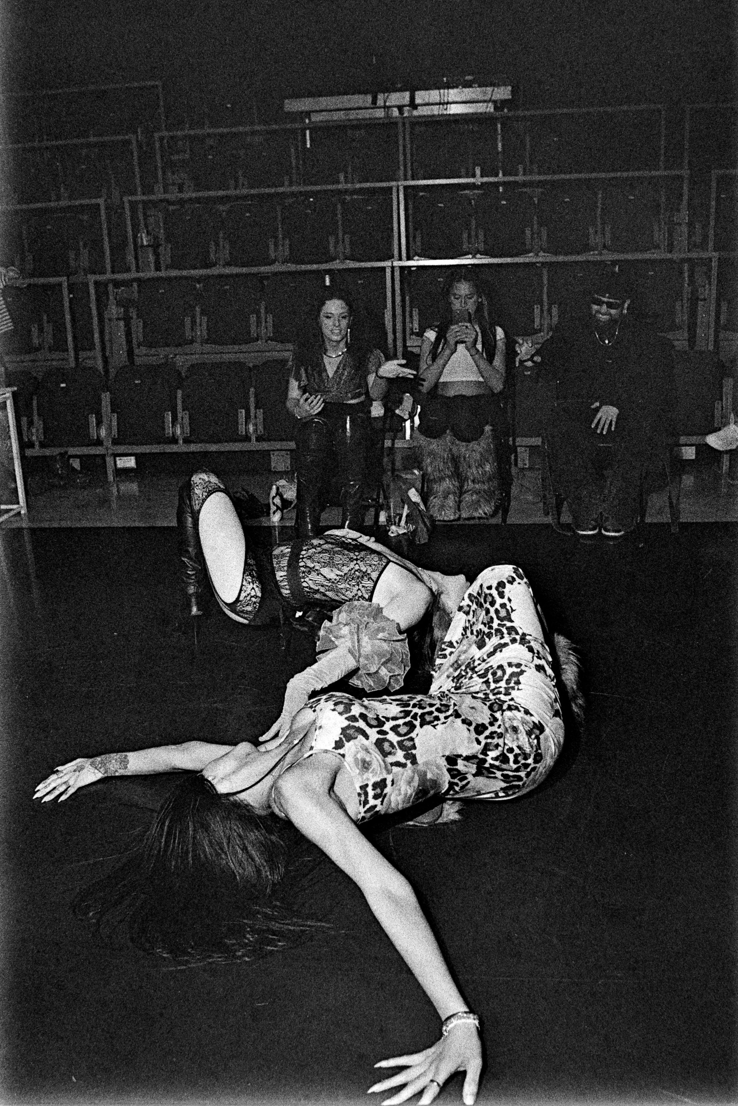A woman wearing leopard print pajamas and high heels is lying on the floor in the foreground with her head and one arm extended outward. In the background, three women are sitting in front of a row of storage lockers; one woman is reading a book, ano
