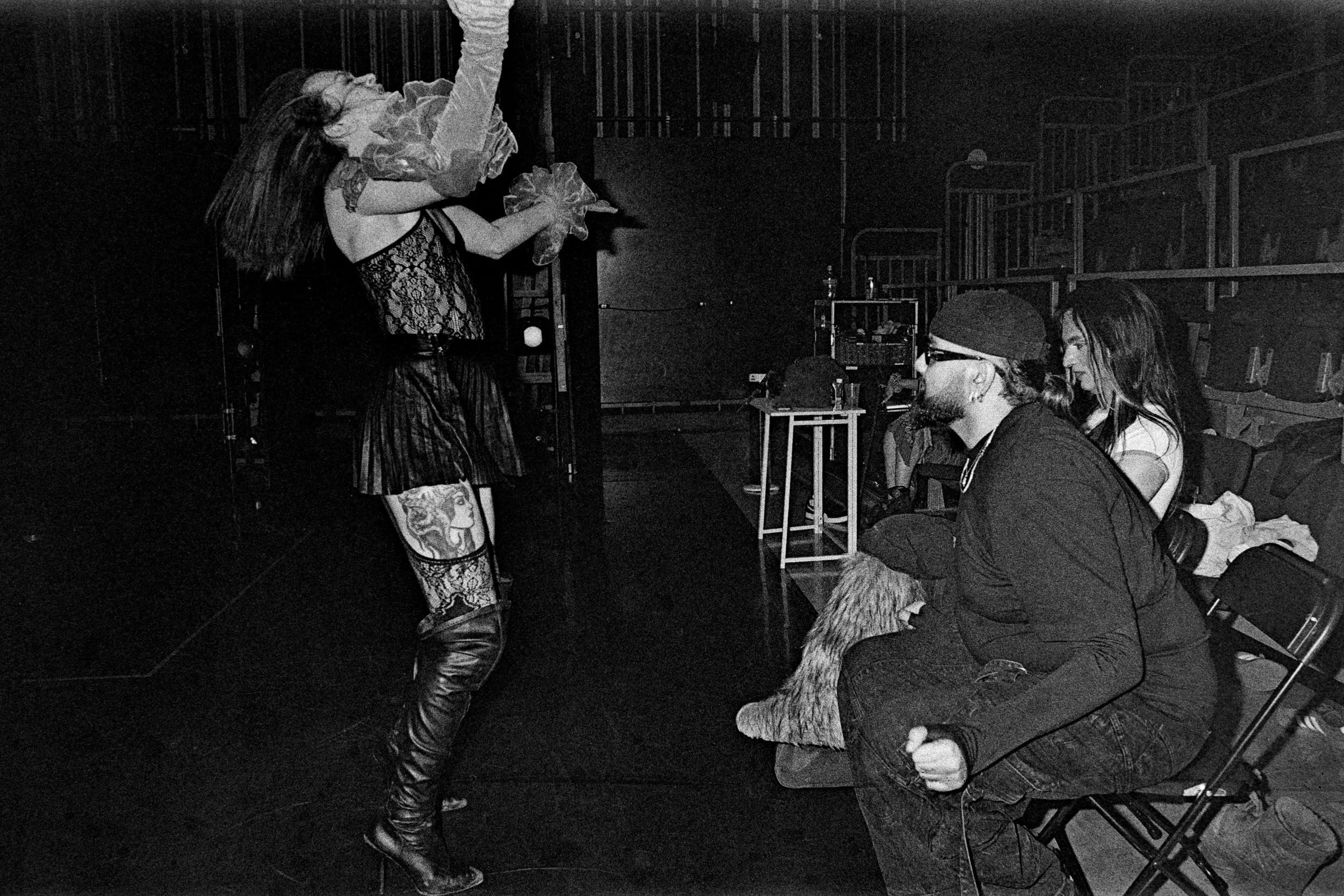 A woman with tattoos on her legs and long hair performing on stage with a microphone, wearing fishnet and lace clothing, while three people in audience seats watch her. The audience members include a person with glasses and a beard, wearing a beanie,