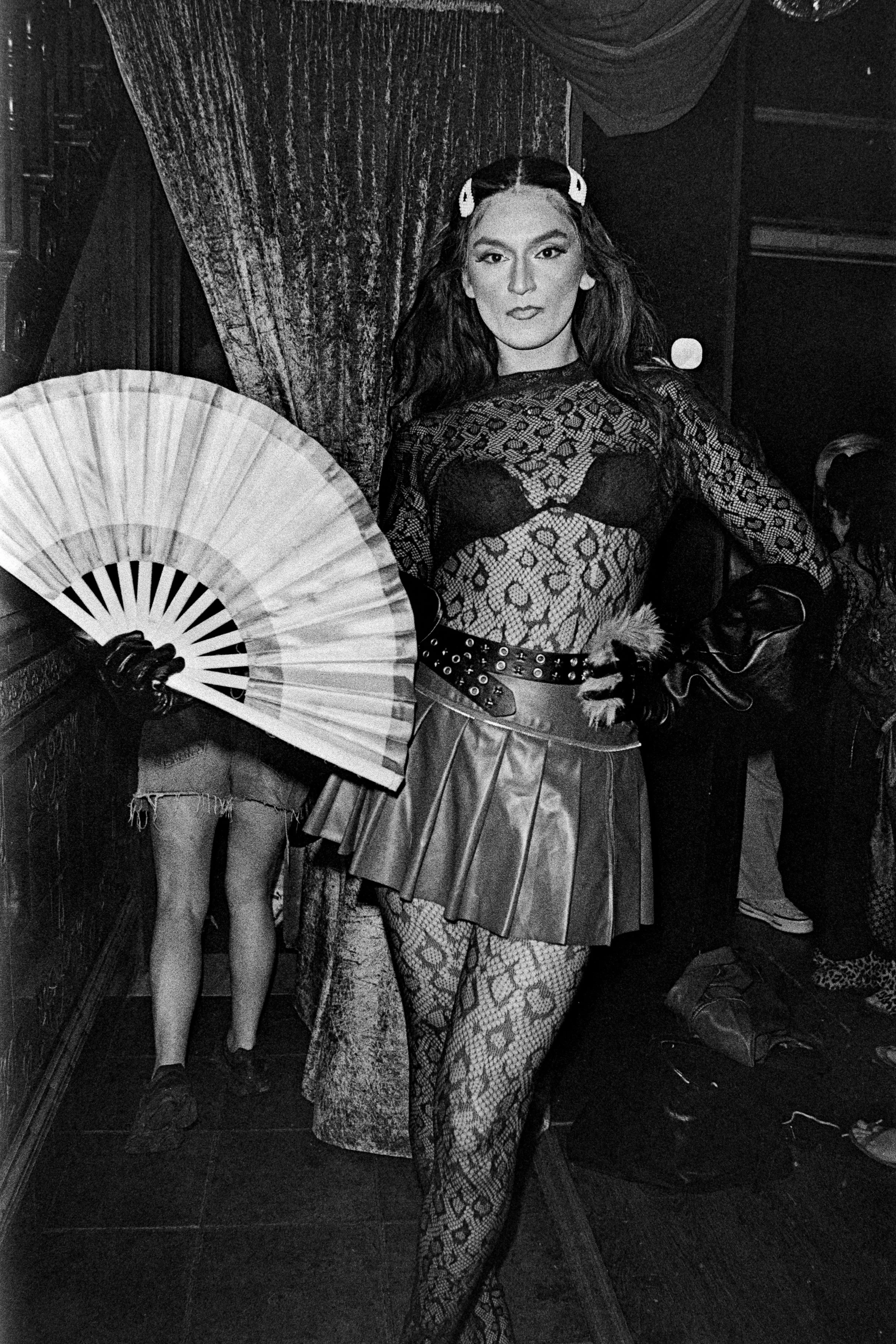 A woman in a fishnet patterned outfit, holding a fan, standing in front of curtains, with another person partially visible in the background.