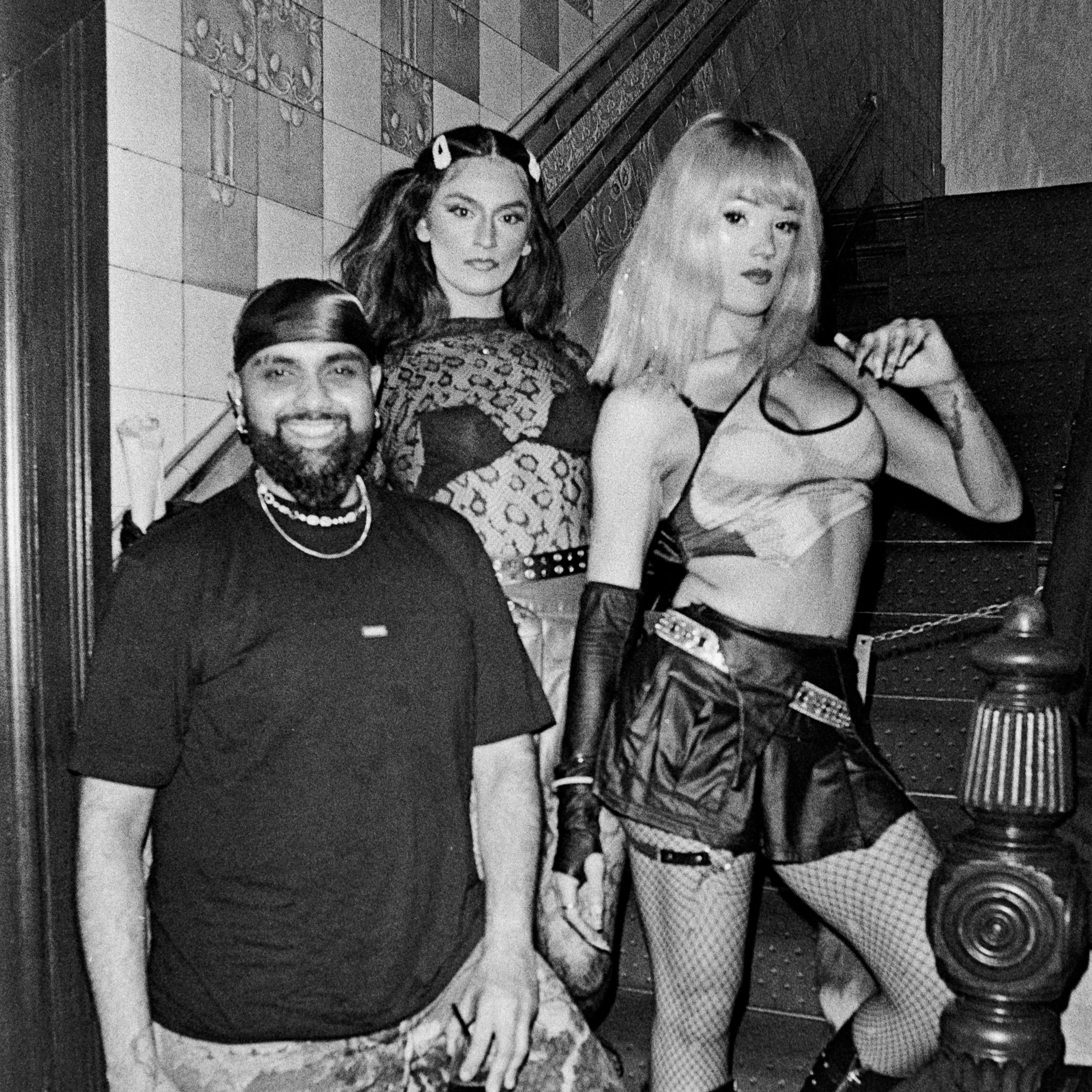 Group of four young people sitting on a staircase, smiling and looking at the camera, dressed in trendy clothing with fishnet stockings and accessories, in an indoor setting with tiled and wallpapered walls.