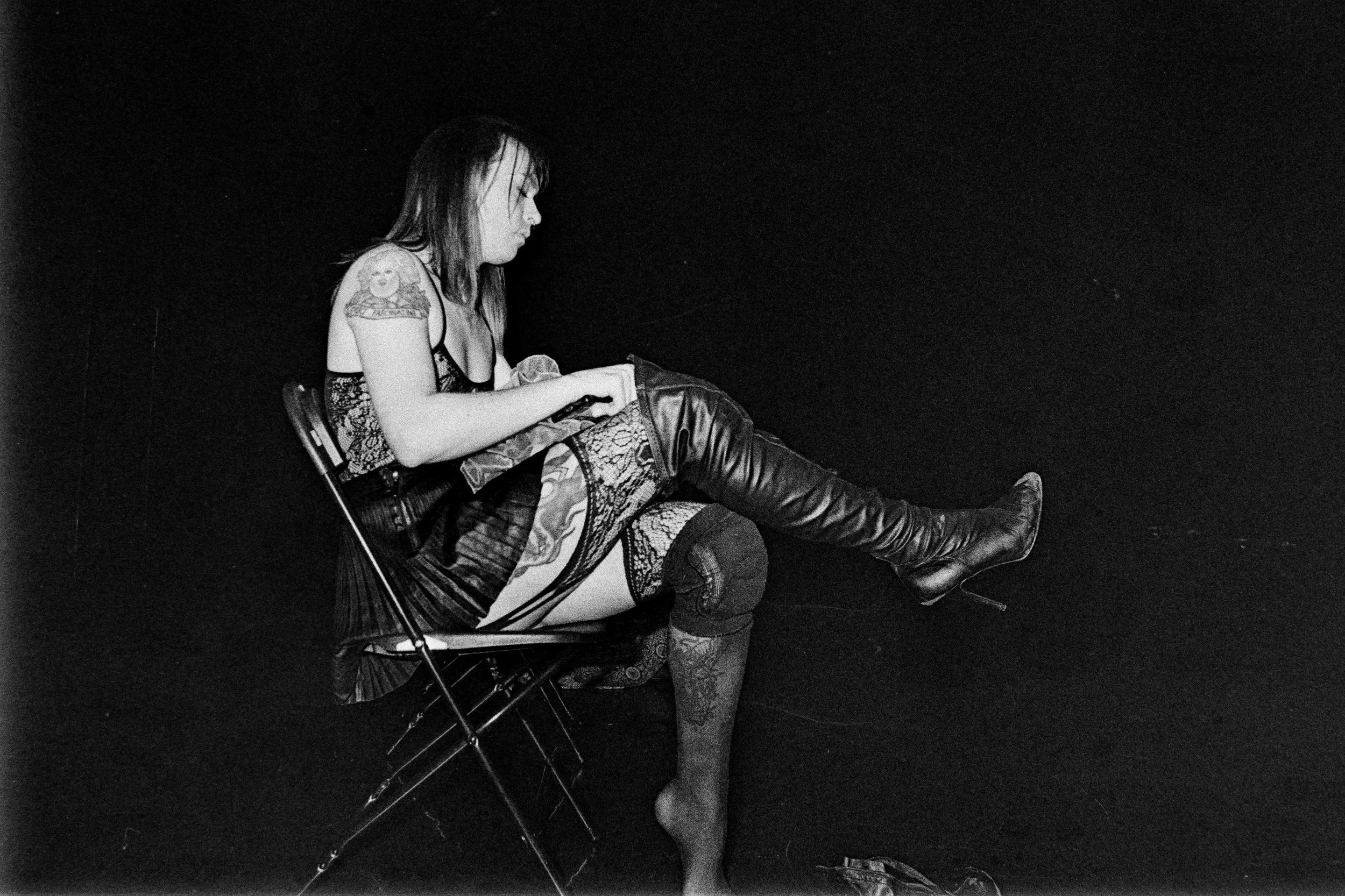 A black and white photograph of a woman sitting on a folding chair against a dark background. She has shoulder-length hair, tattoos on her arm, and is wearing a patterned dress, thigh-high boots, and lace stockings.