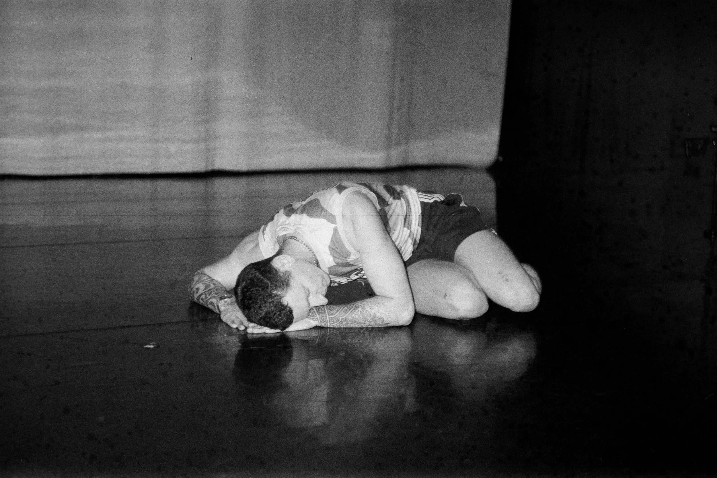 A black and white photo of a person lying on the floor, with their head resting on their arm and eyes closed, dressed in casual clothing, next to a wall and a curtain.