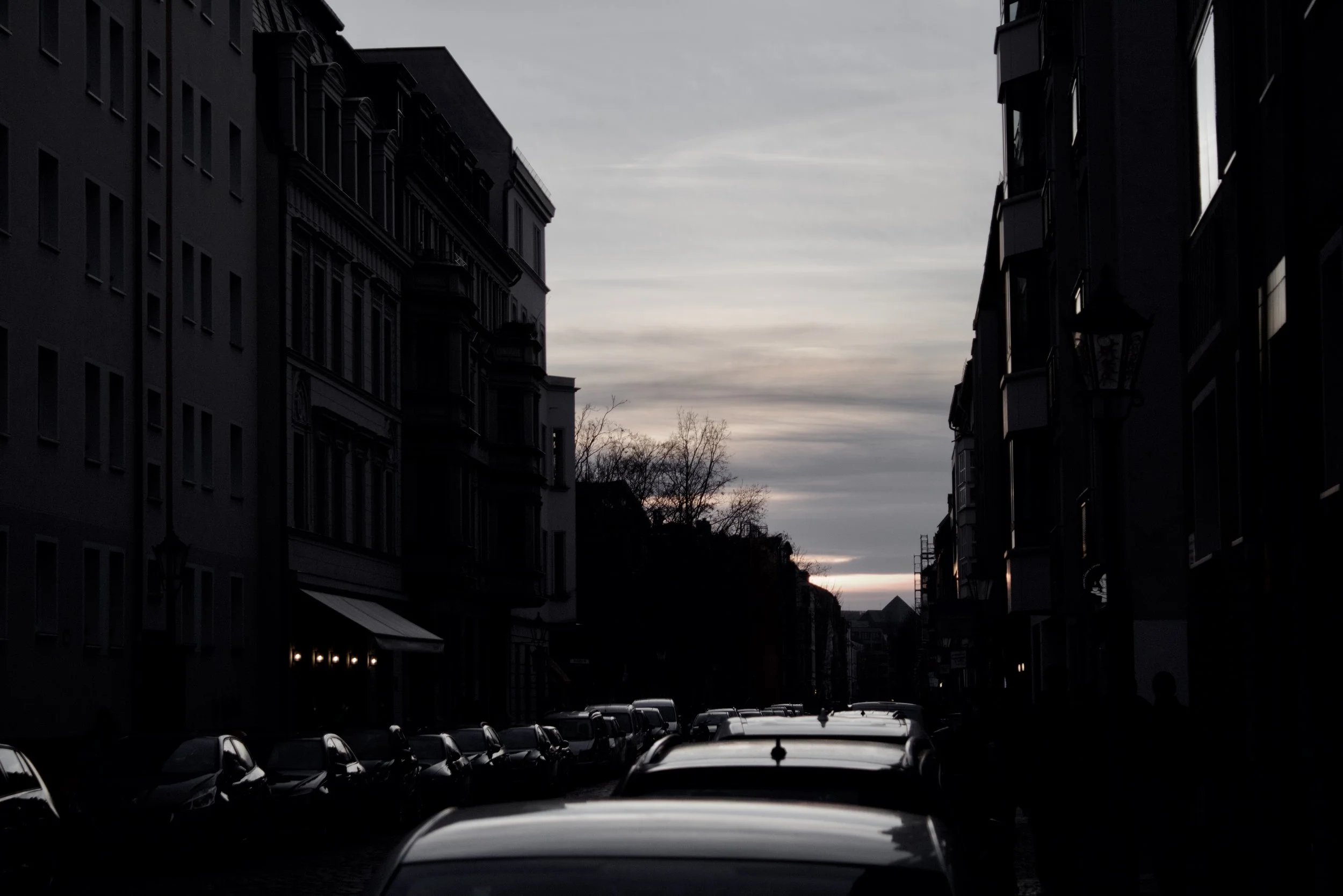 A street scene at dusk with cars parked on both sides and tall buildings lining the street. The sky has a mix of light and dark clouds.
