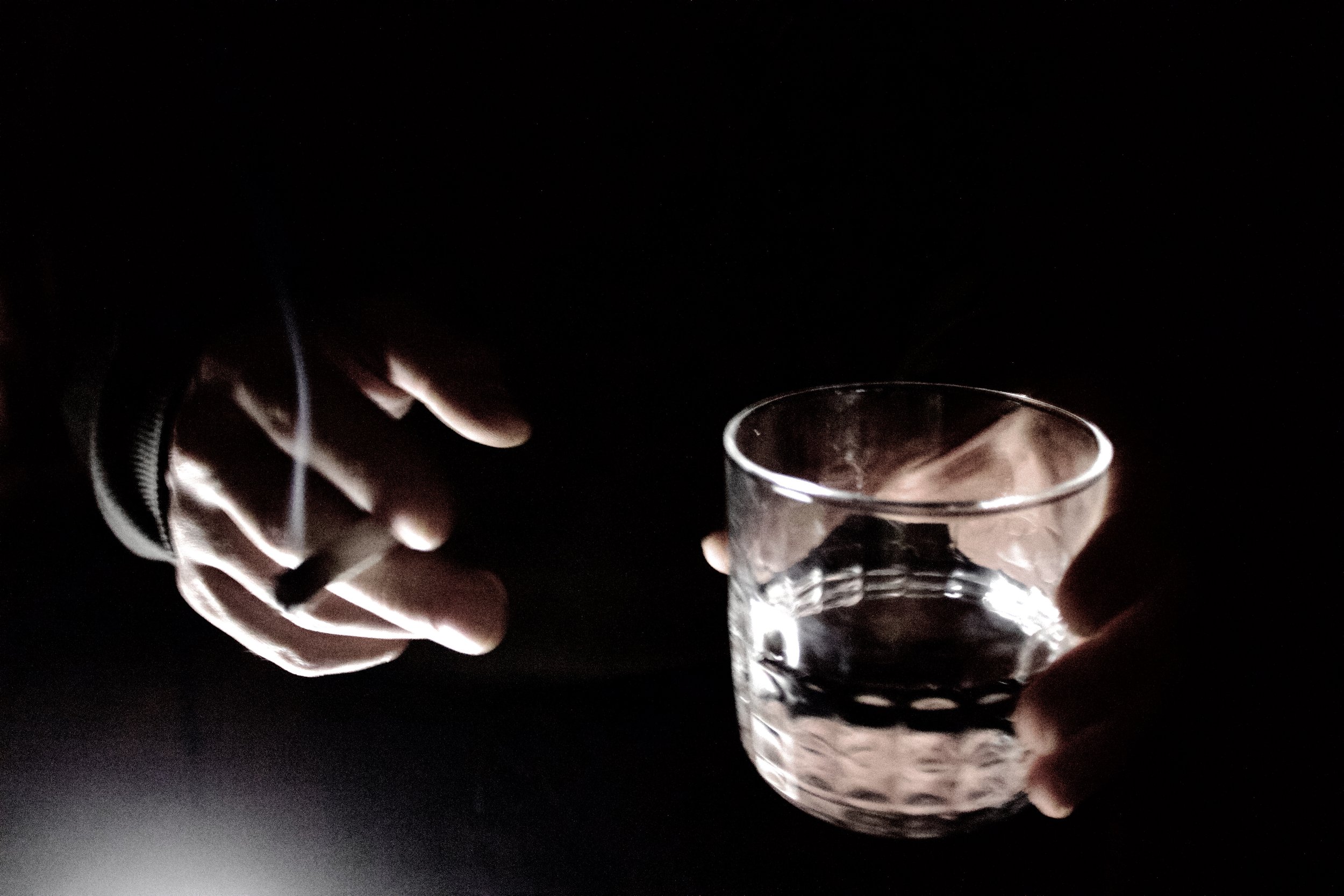 A hand holding a clear glass of water with dark objects inside, against a dark background. The wrist is partially covered by a jacket.