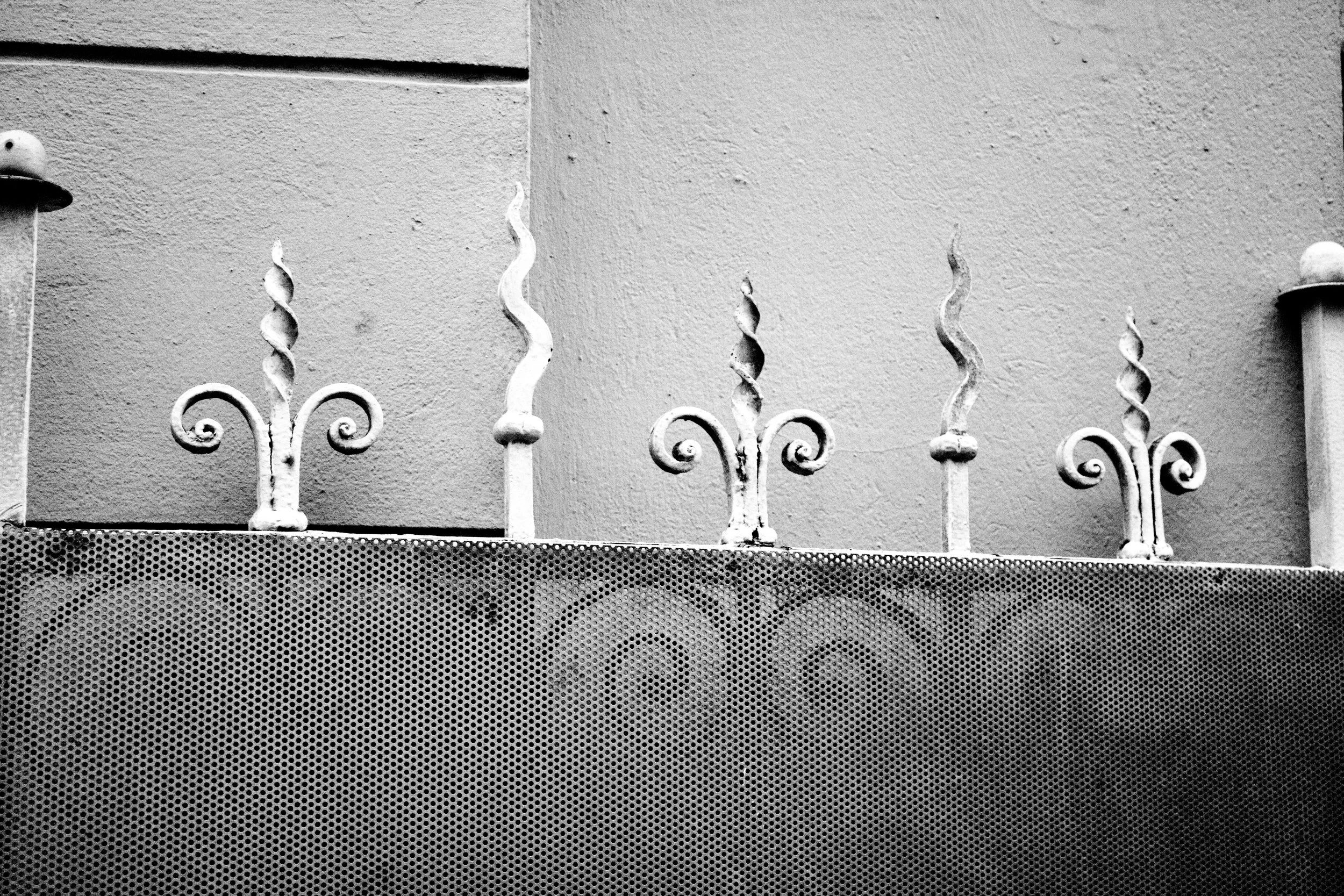 Black and white photo of a decorative metal fence with ornate, curly designs, set against a textured wall, with a perforated metal sheet in the foreground.