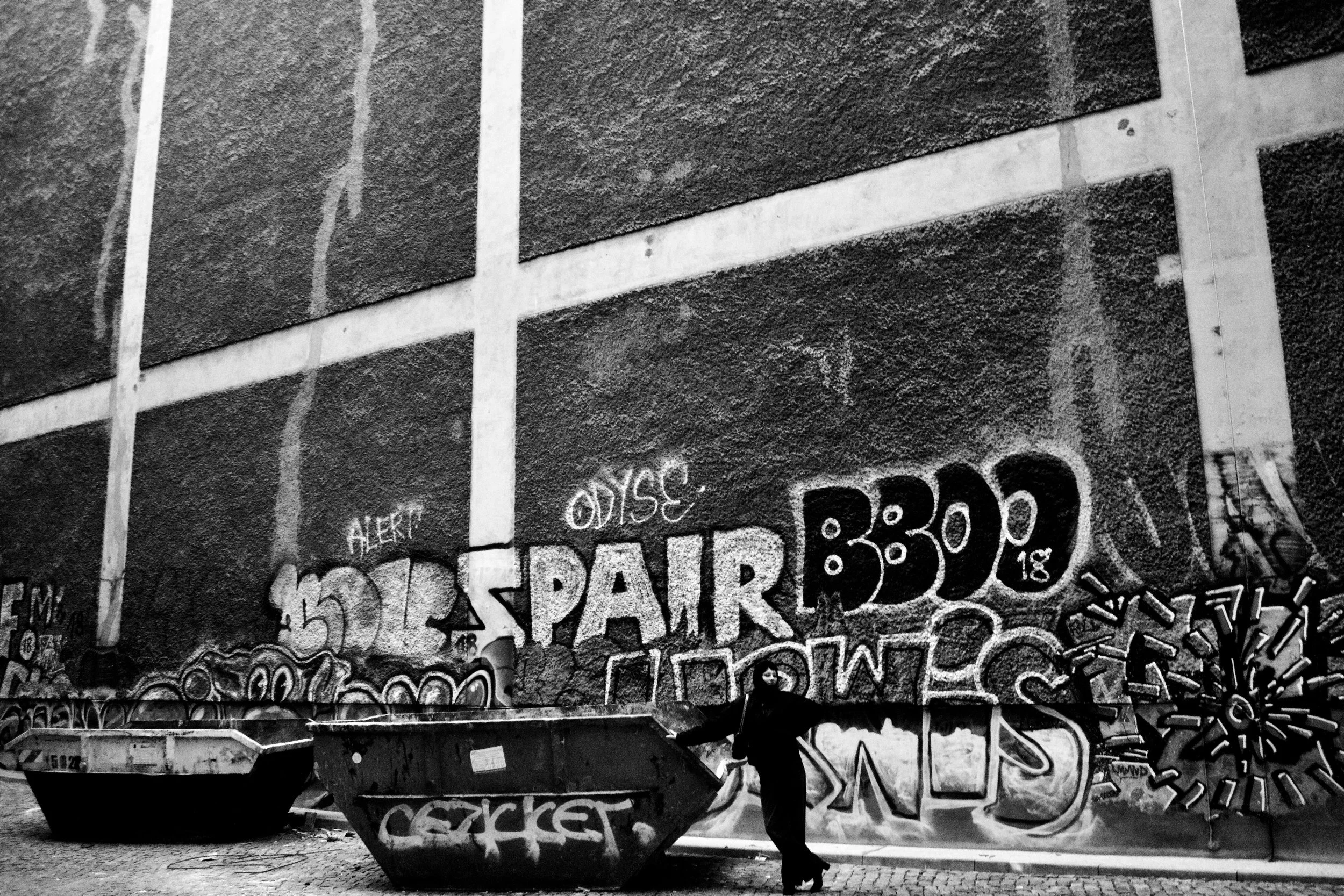 Black and white photo of a building wall covered in graffiti, with a person standing under an umbrella and a boat parked in front.