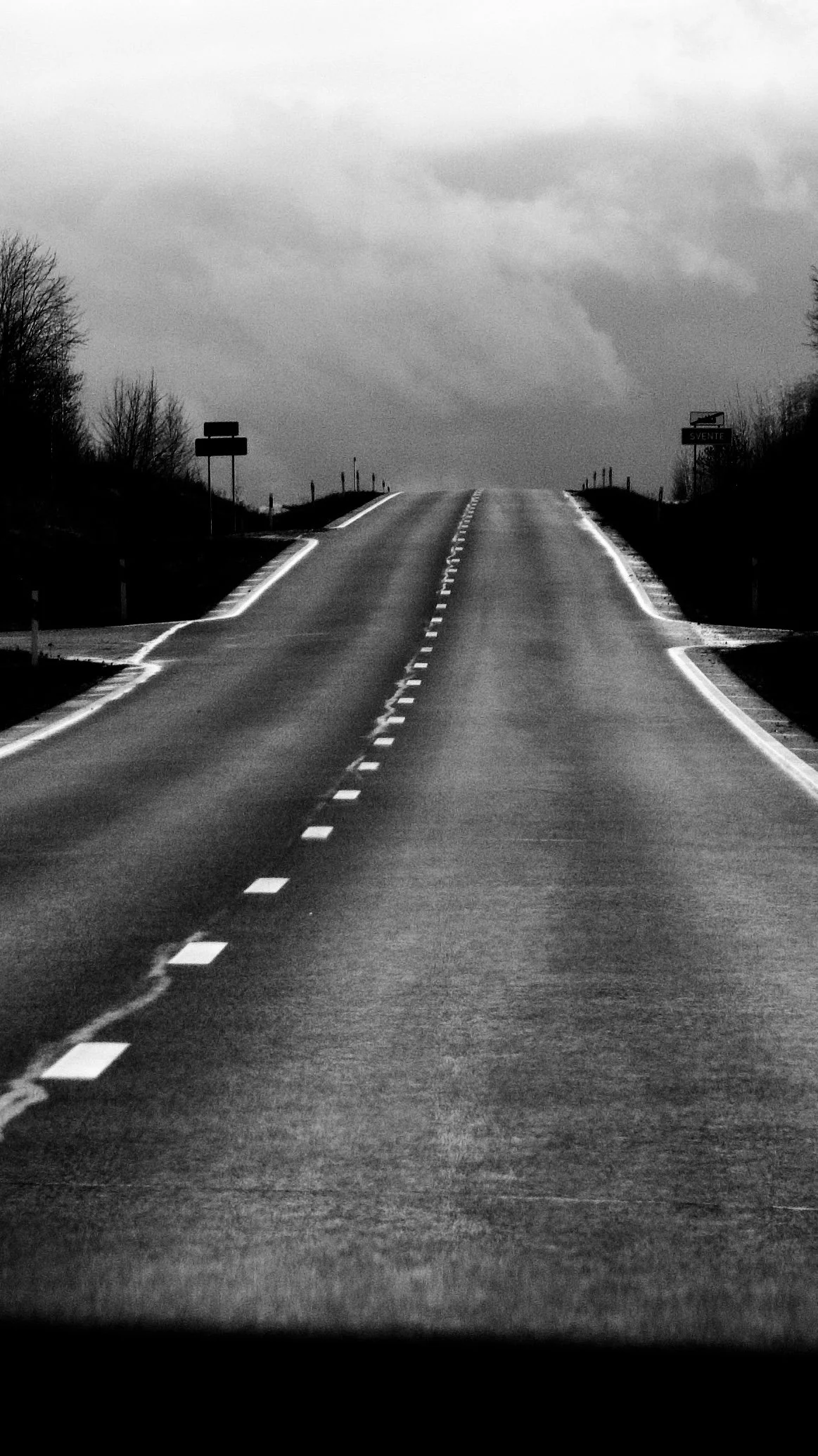 Black and white photo of an empty winding road with trees on either side and cloudy sky overhead.